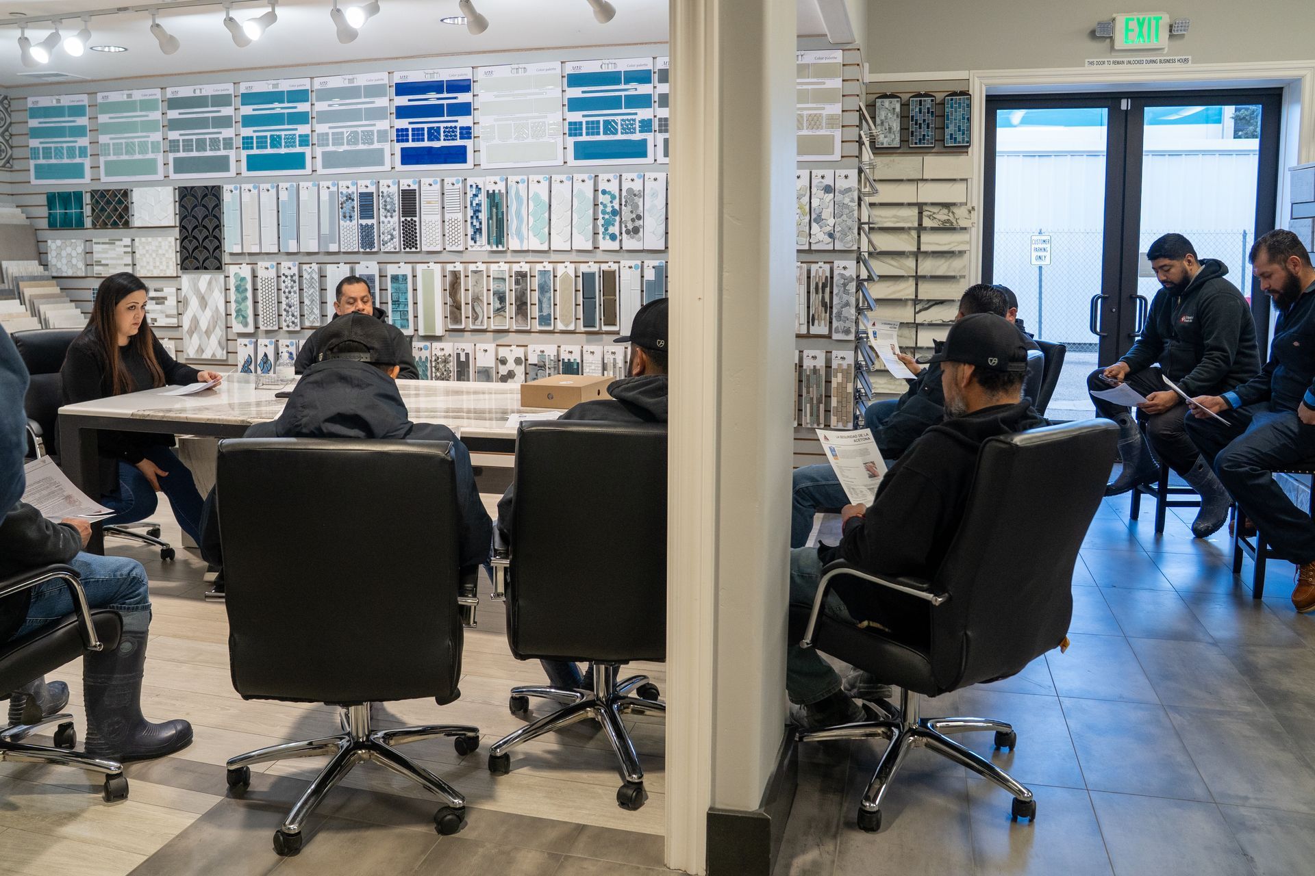 A group of people in a showroom, some seated at a table, others in a circle. Tiles displayed on walls.