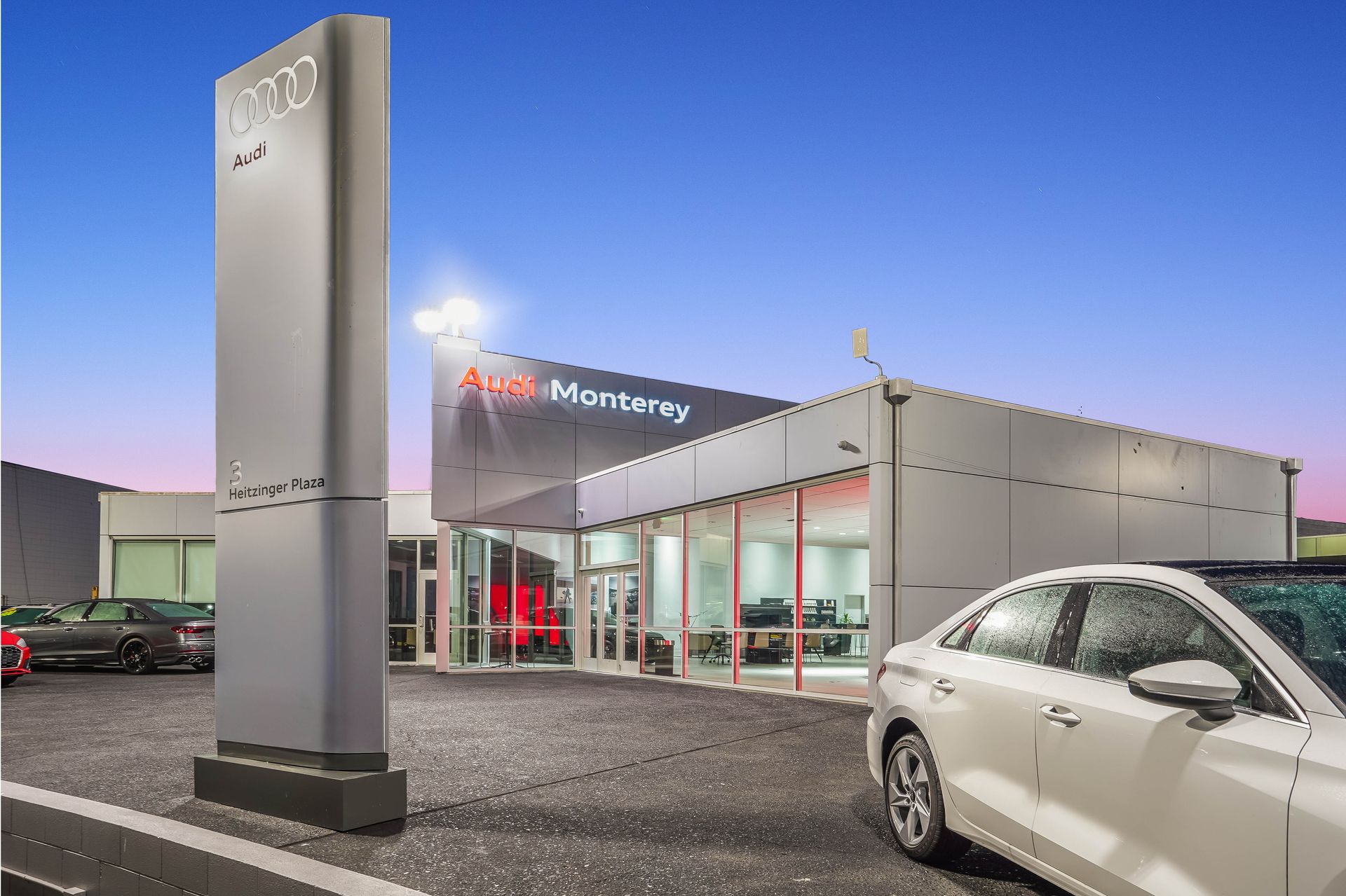 Audi Monterey dealership exterior at dusk, sign, modern building, white sedan in foreground.