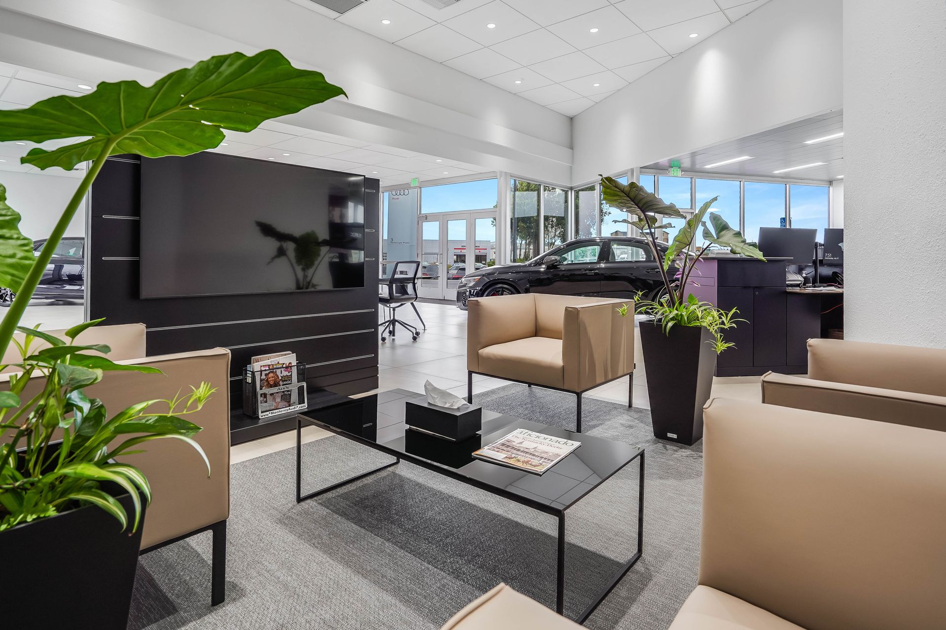 A modern waiting area with seating, large screen, plants, and windows overlooking a car.