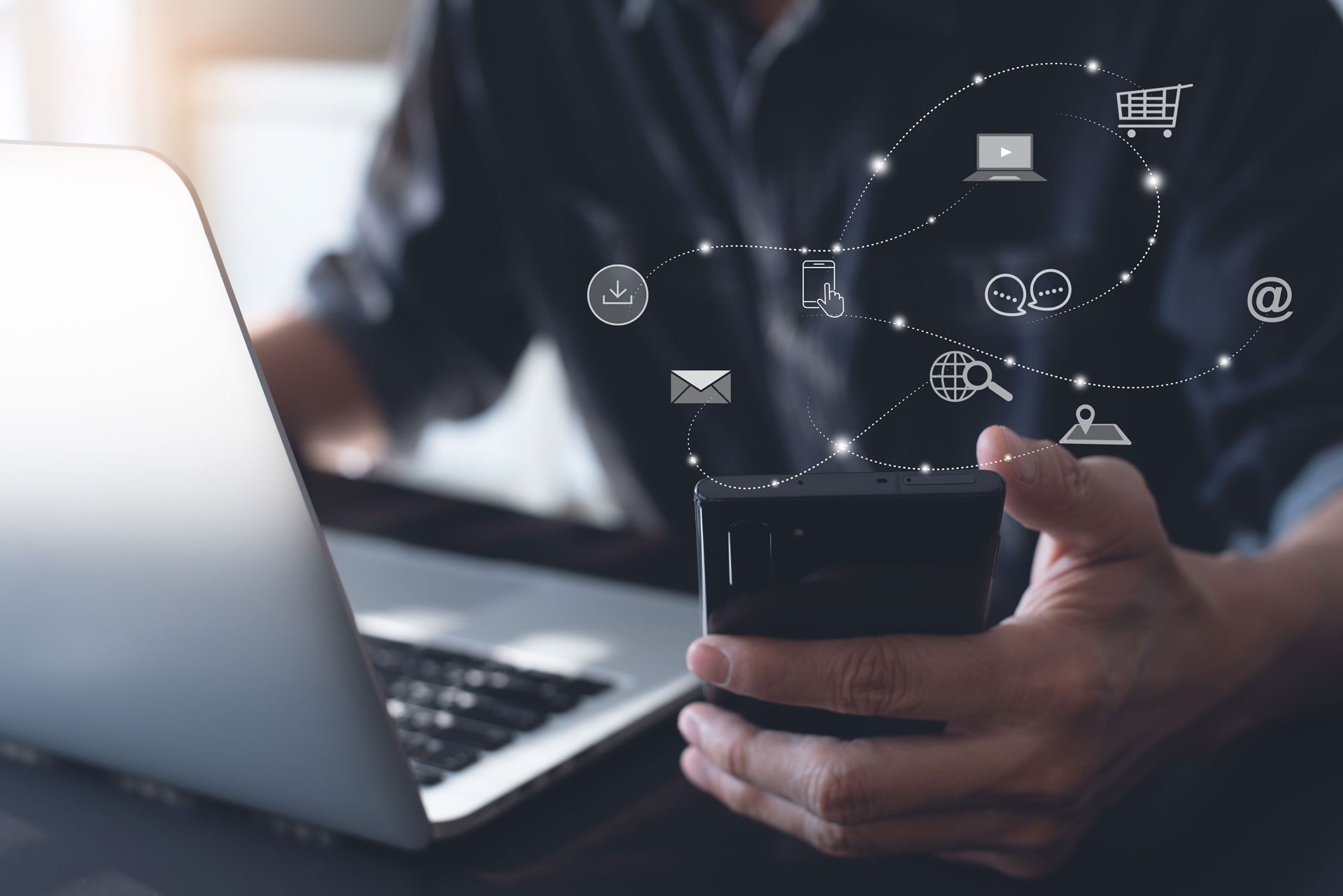 A man is holding a cell phone in front of a laptop computer.