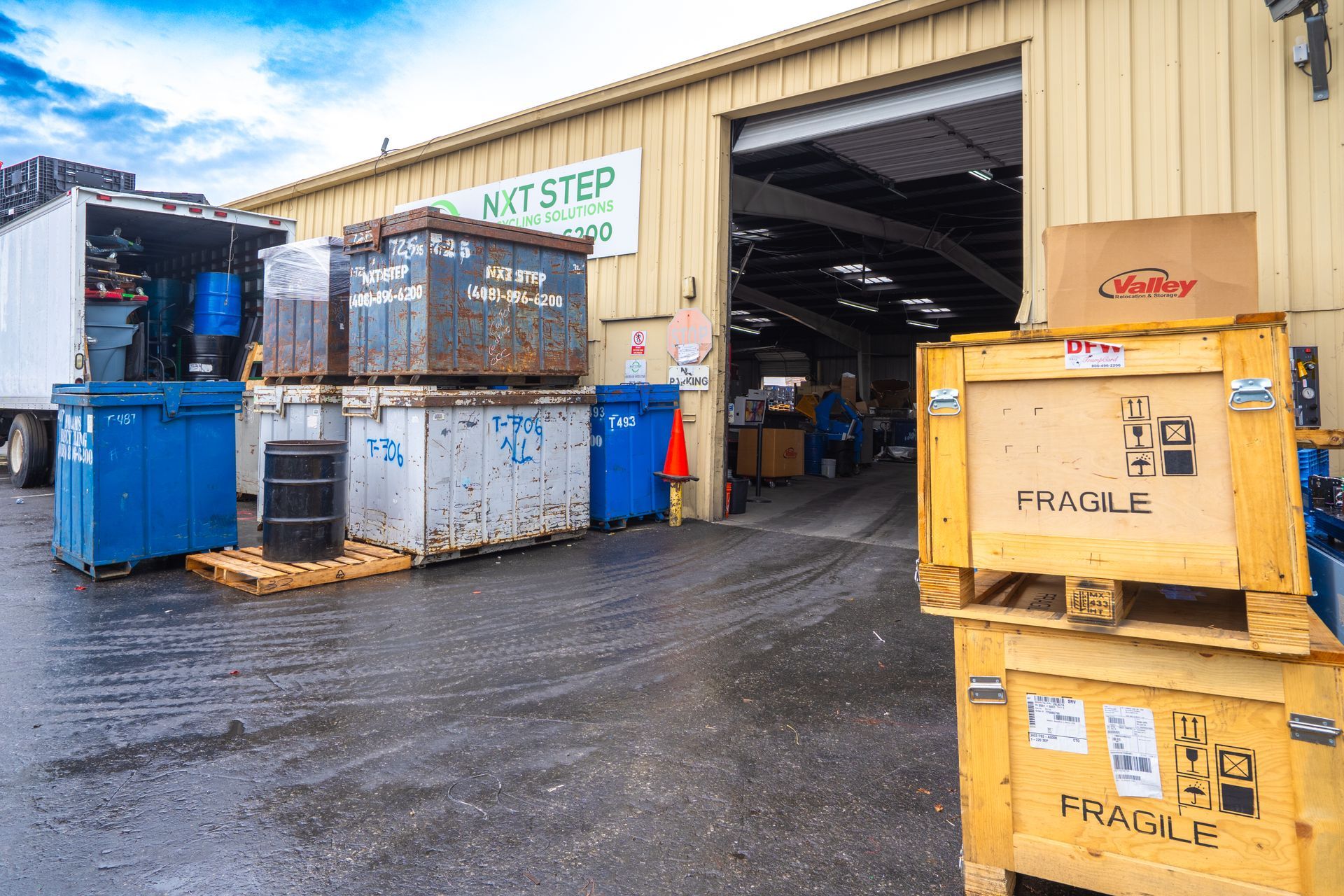 Exterior of a warehouse with stacked wooden crates, dumpsters, and a semi-truck under a cloudy sky.
