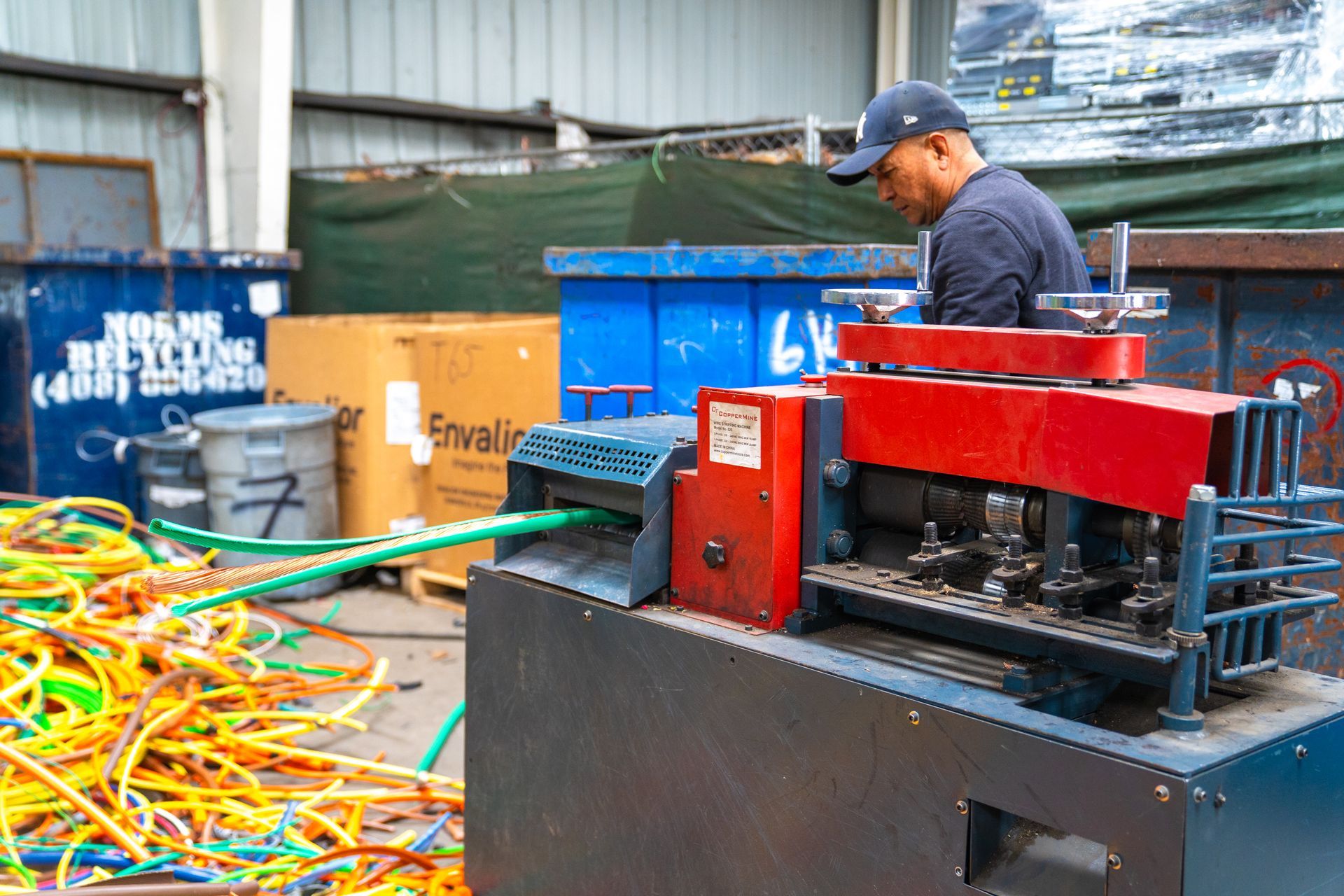 Man operating a machine to strip wires for recycling; outdoors, blue bins, green and yellow wires.