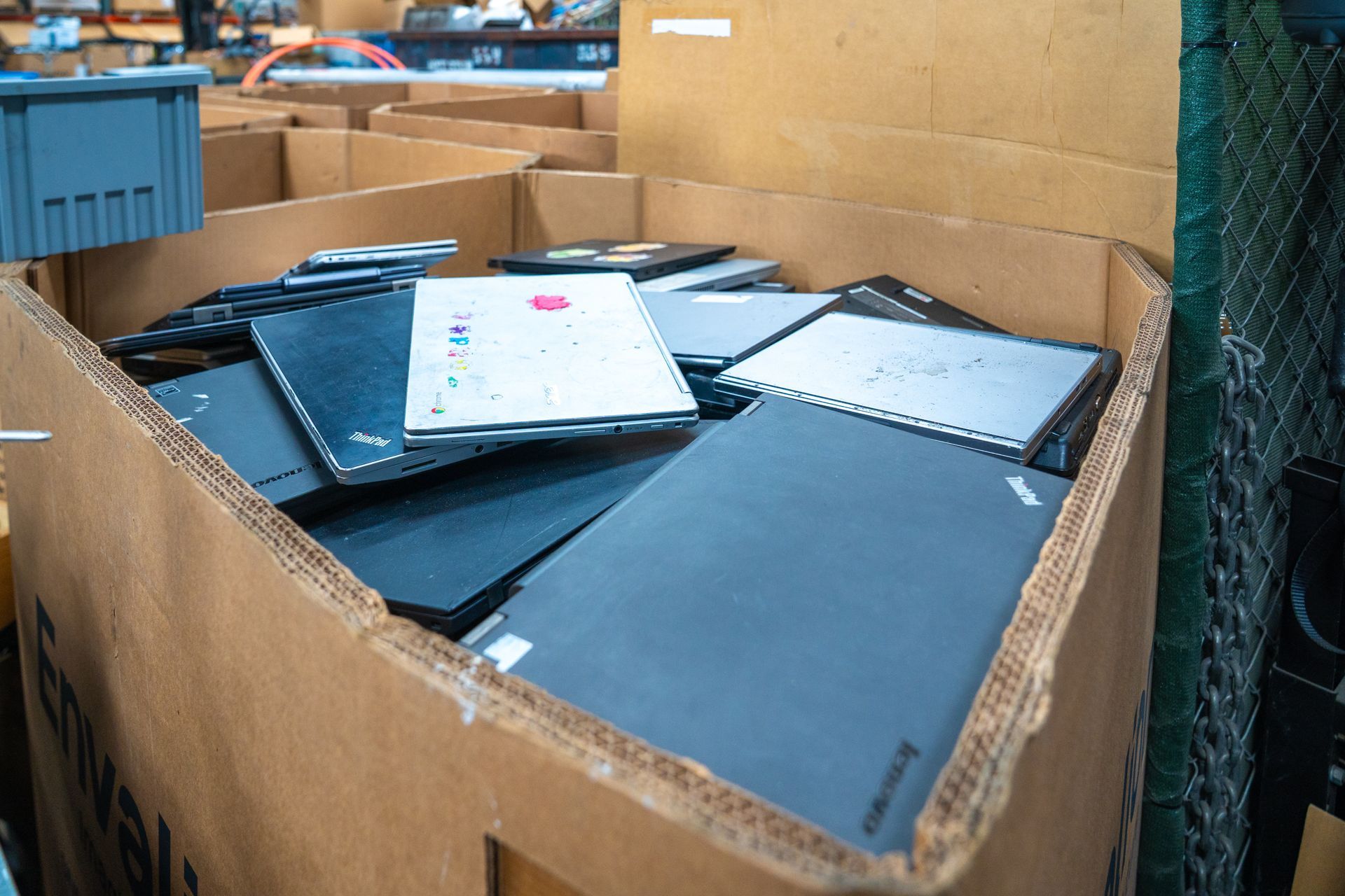 Cardboard box overflowing with closed laptops in a warehouse setting.
