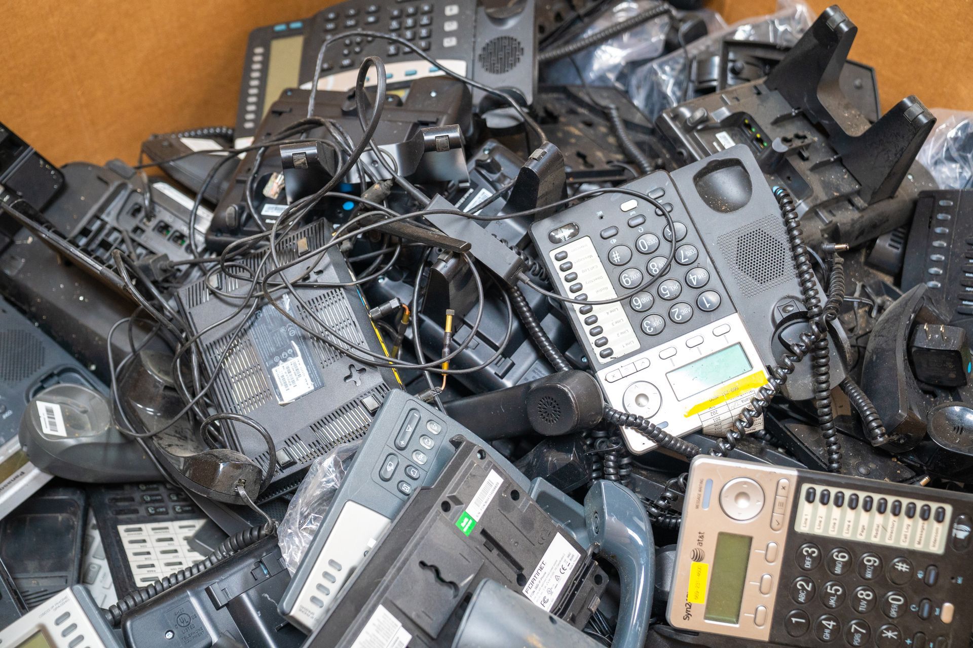 Pile of discarded black office phones and tangled cords in a cardboard box.