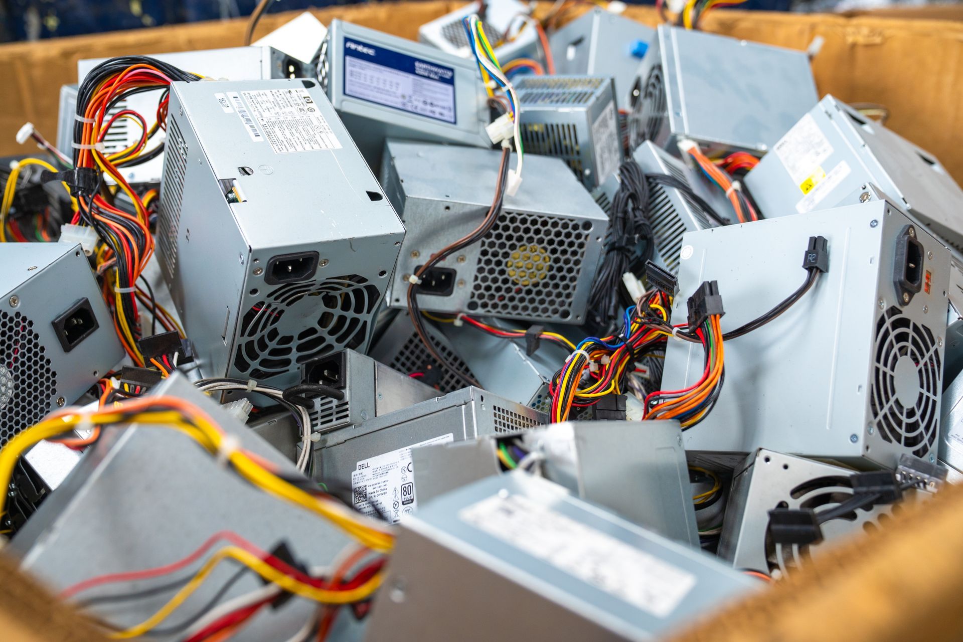 Cardboard box overflowing with gray computer power supply units and tangled colored wires.