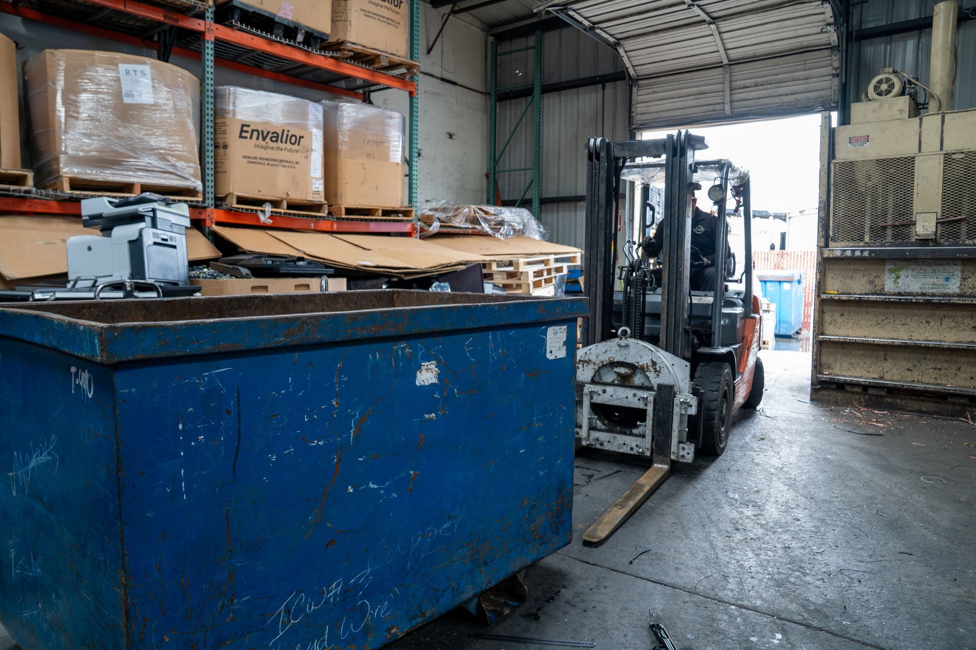 Forklift loading a blue dumpster in a warehouse with boxes on shelves.