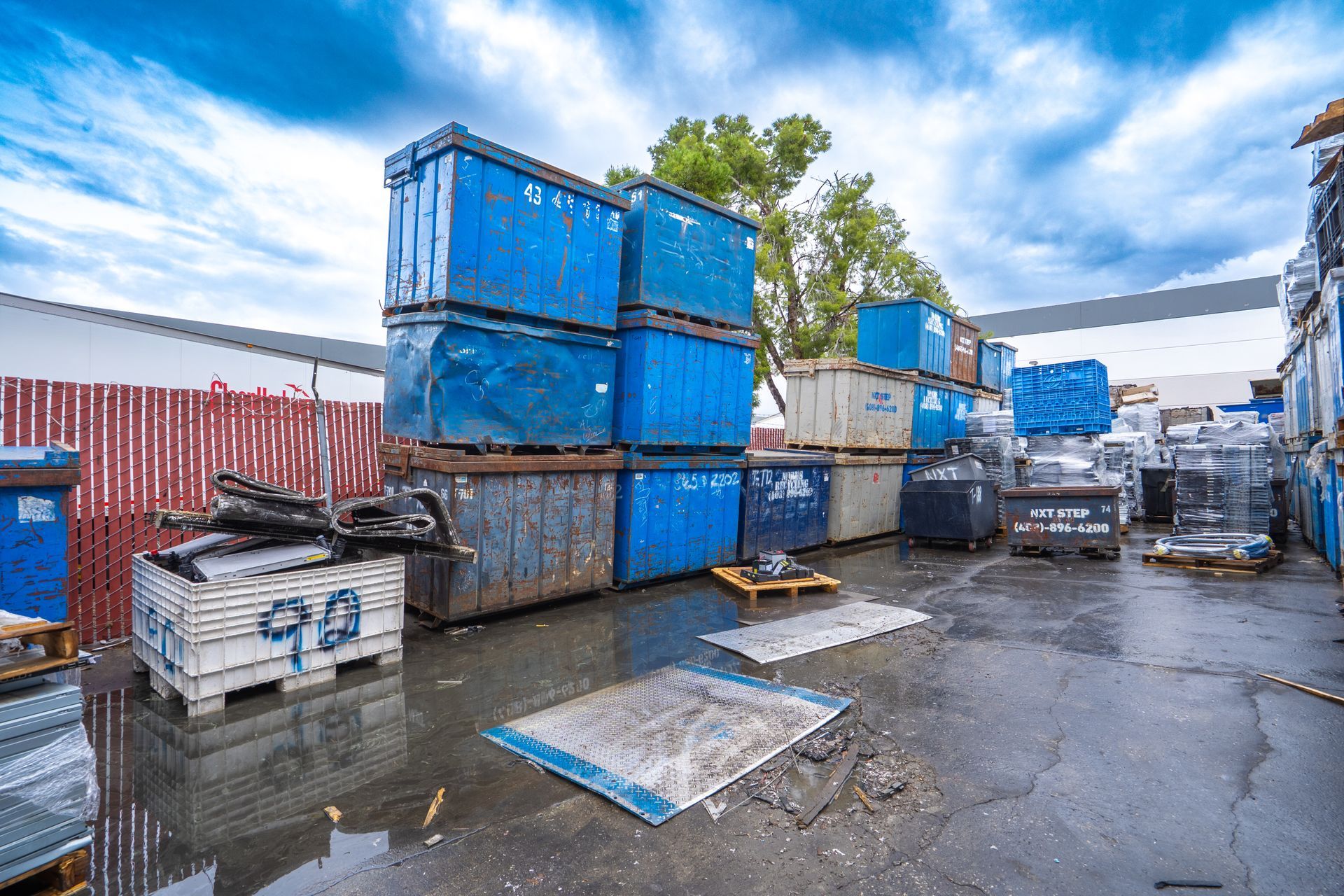 Blue dumpsters stacked in a lot, with other containers and a fence, under a cloudy sky.