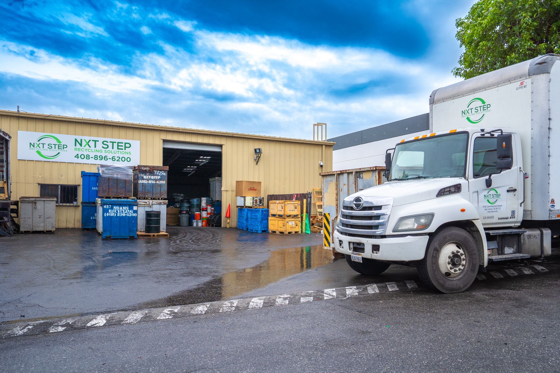 White truck parked outside a warehouse with recycling bins, cloudy blue sky.