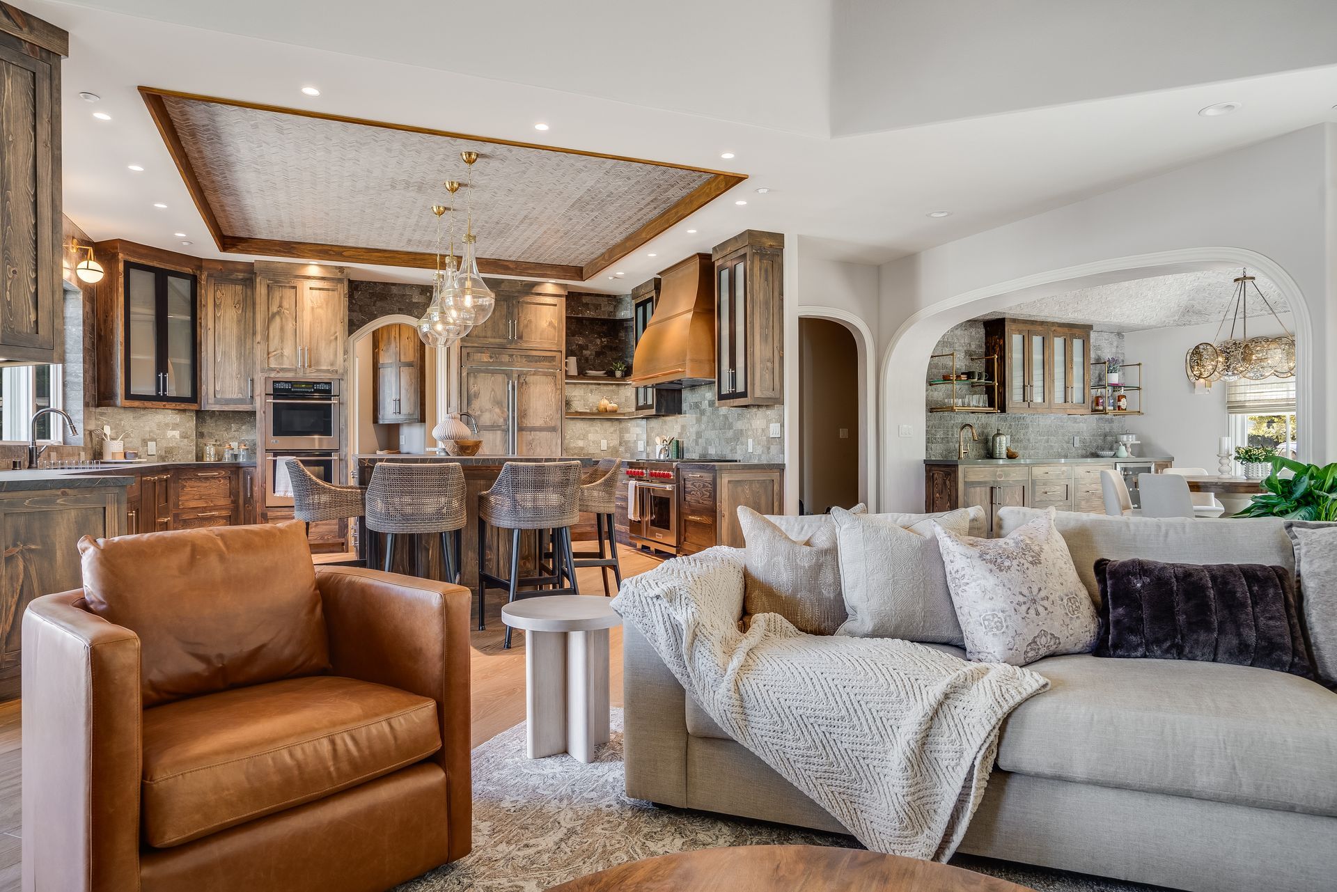 Open-concept kitchen and living area with brown cabinetry, beige couch, leather armchair, and arched entryway.