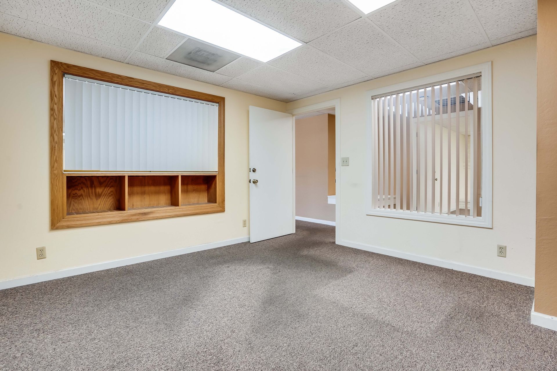 Empty office room with beige walls, carpet, and window blinds; a door leads to another room.