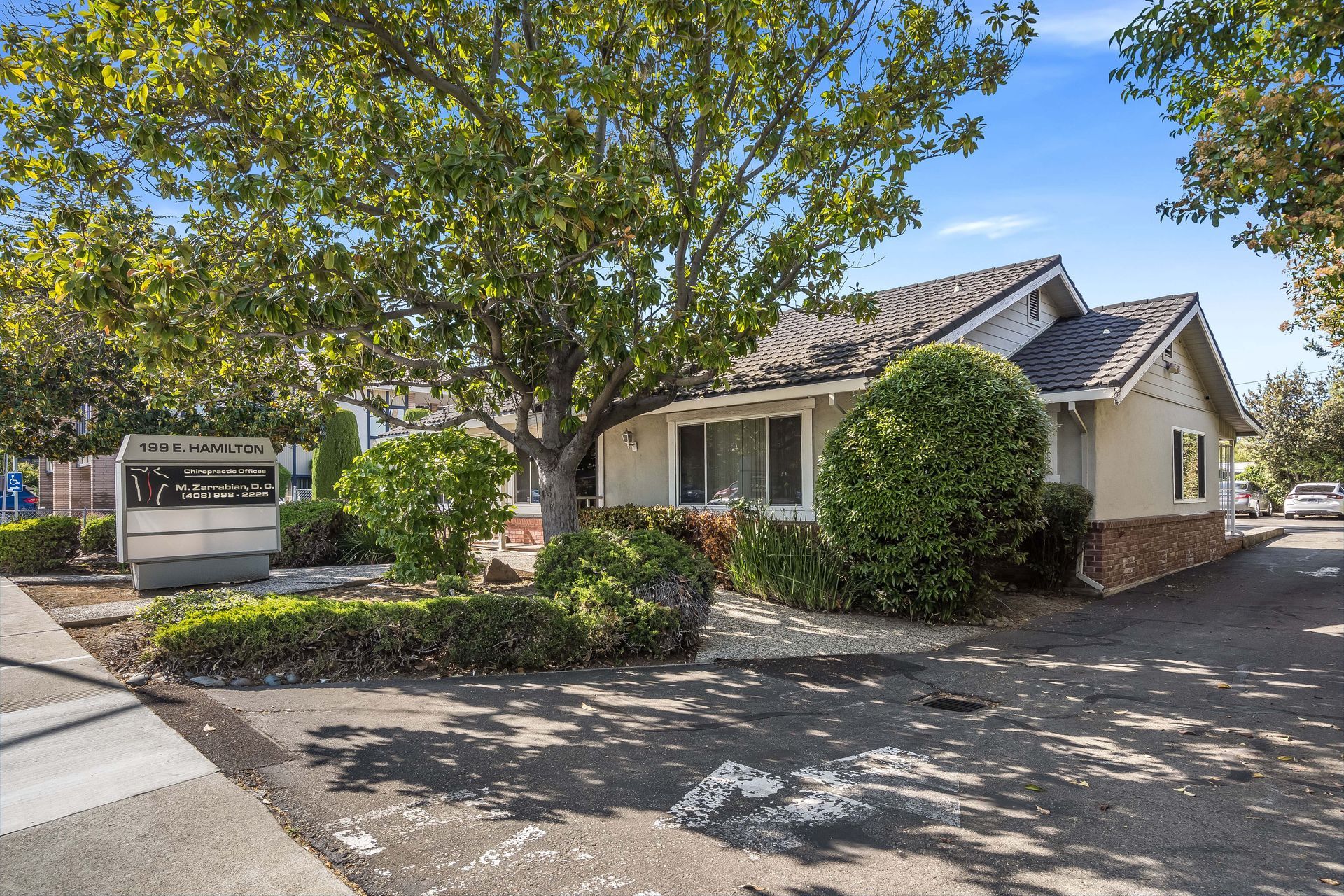 Small beige building with a brick facade, trees in front, and a sign.