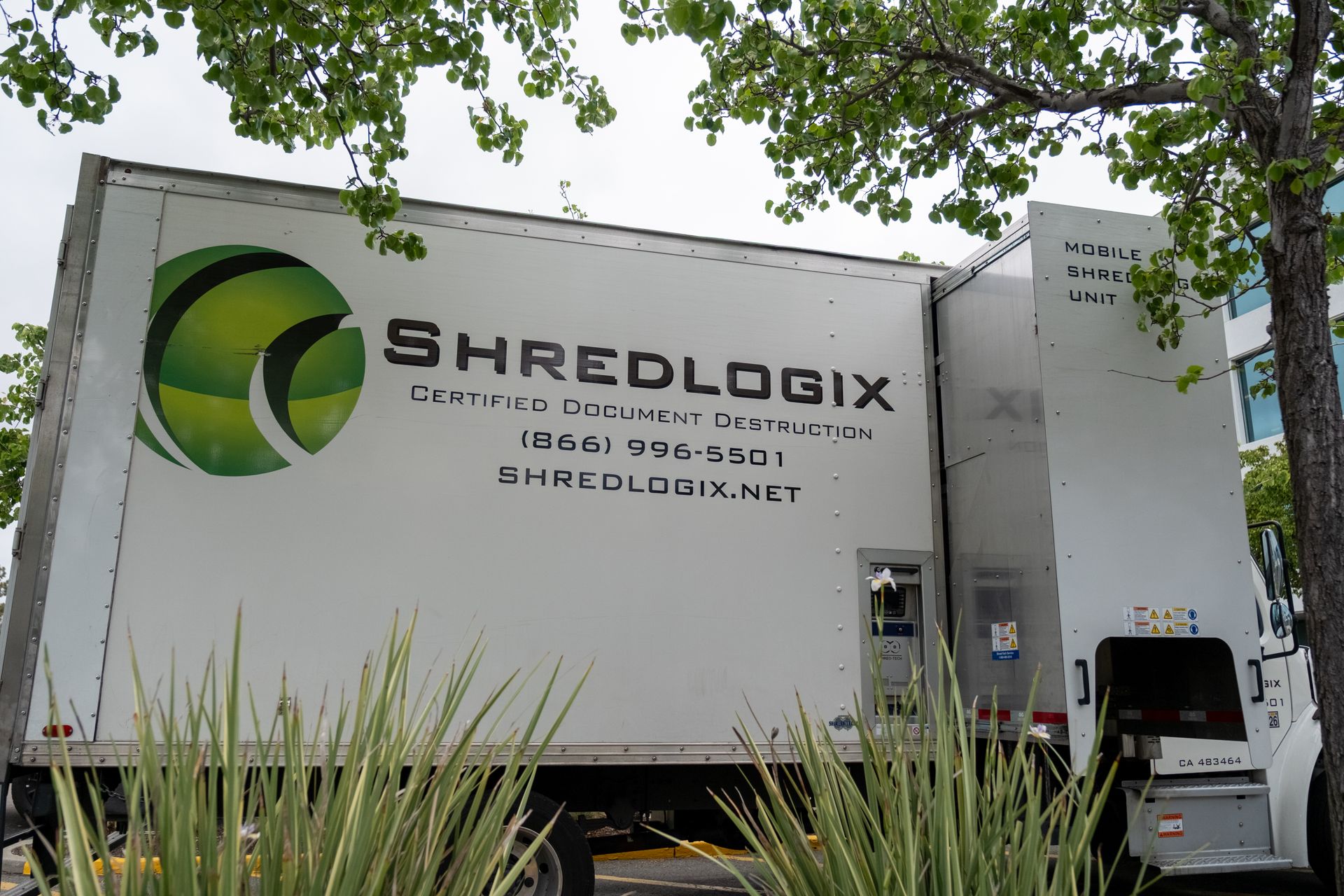 White Shredlogix truck with logo parked outdoors near green plants and trees.