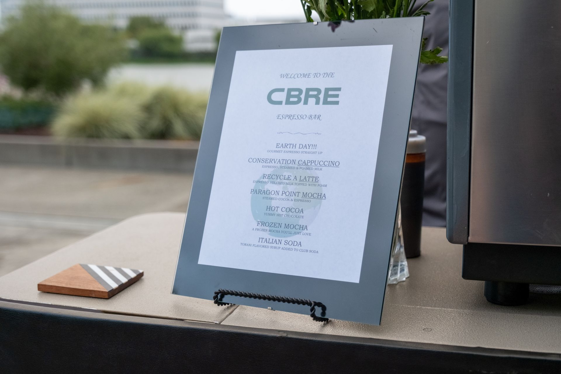 A sign for CBRE on a table with a wooden coaster and a stainless steel appliance outside.