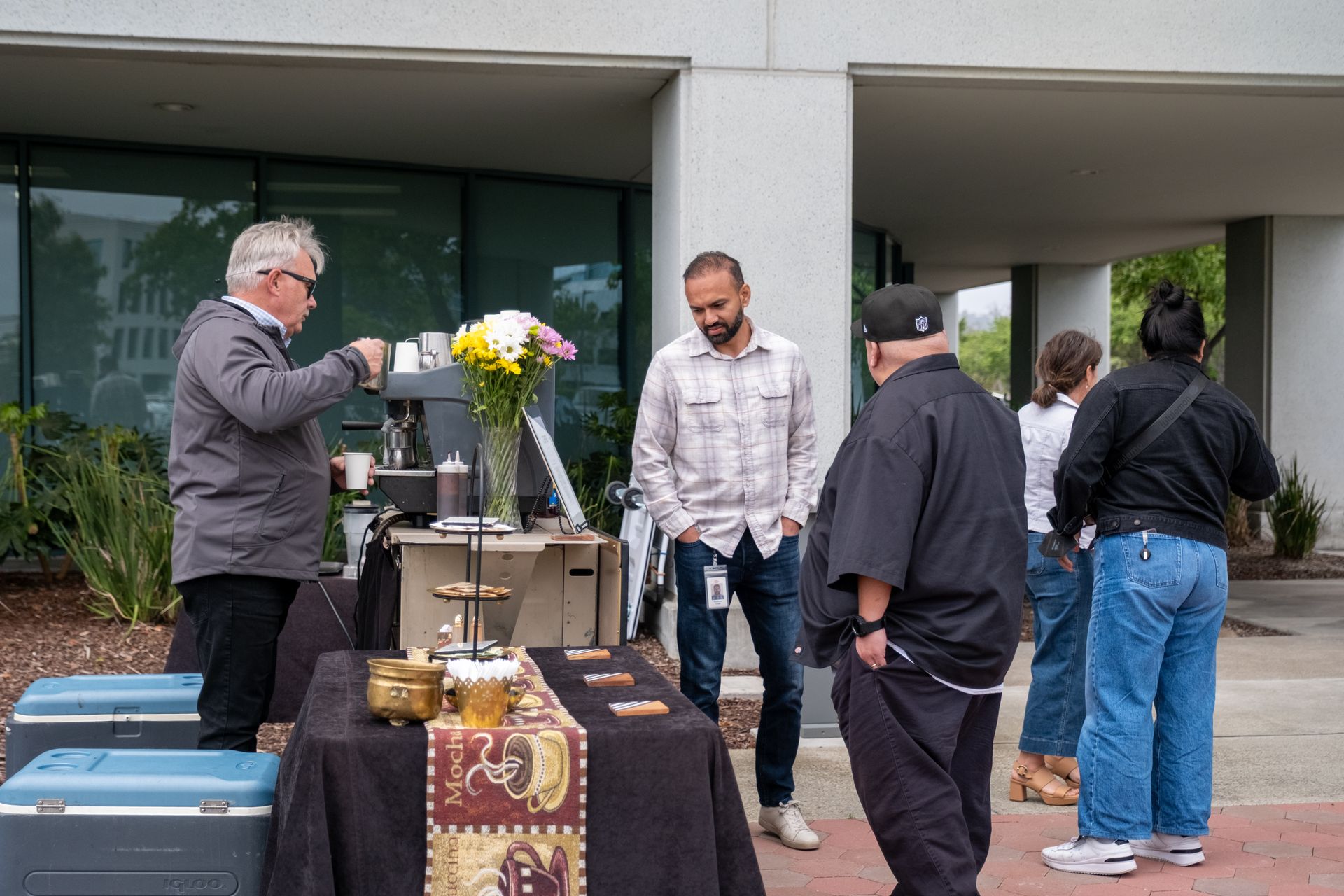 Outdoor coffee station with people; one pours coffee, others wait. Flowers and snacks on table.