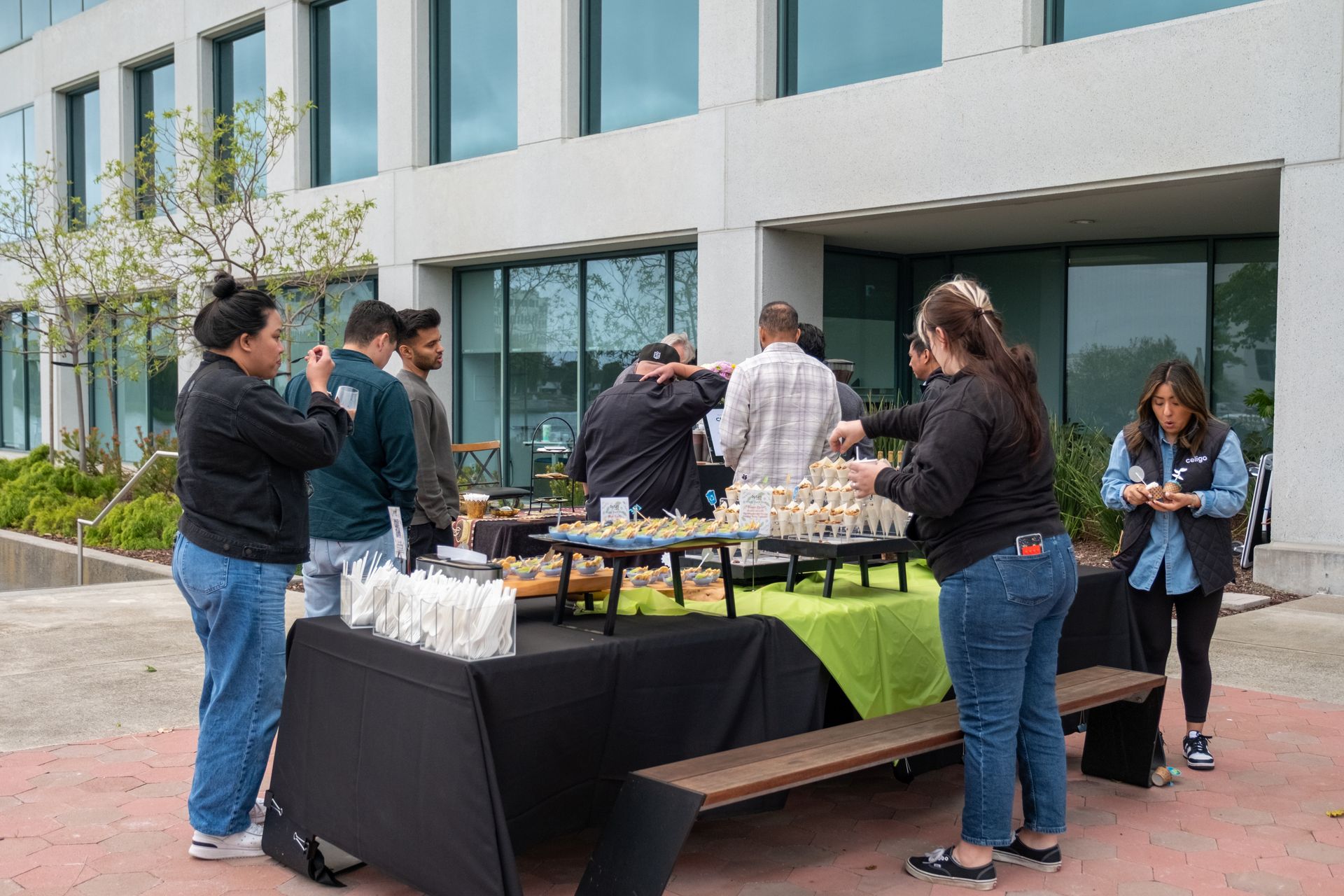 People at an outdoor food table in front of a modern building. Black tablecloth, green accents, food offerings.