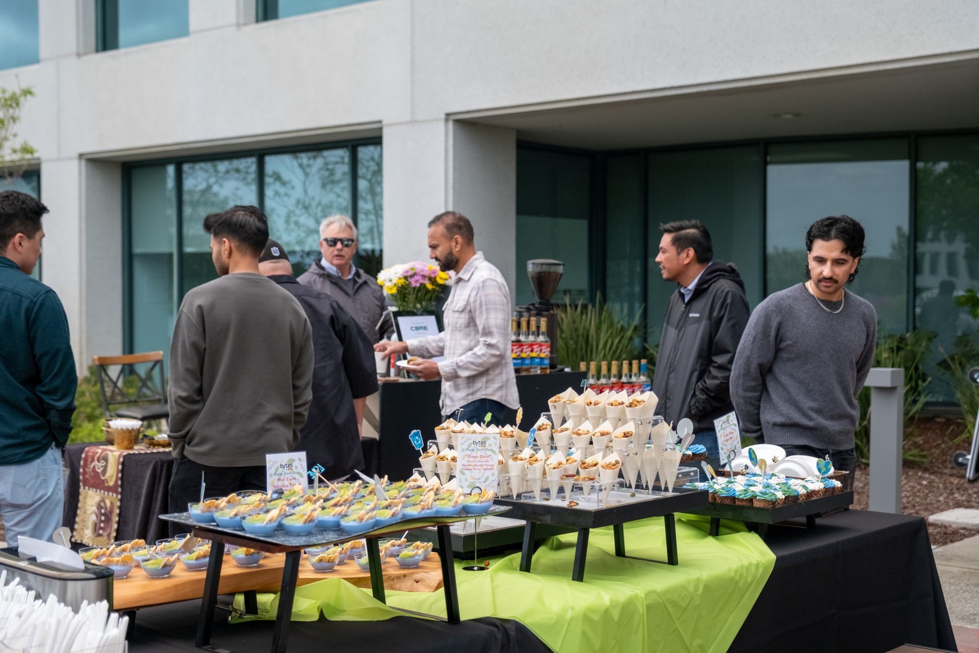 People gathered at an outdoor food table with appetizers and drinks near a modern building.