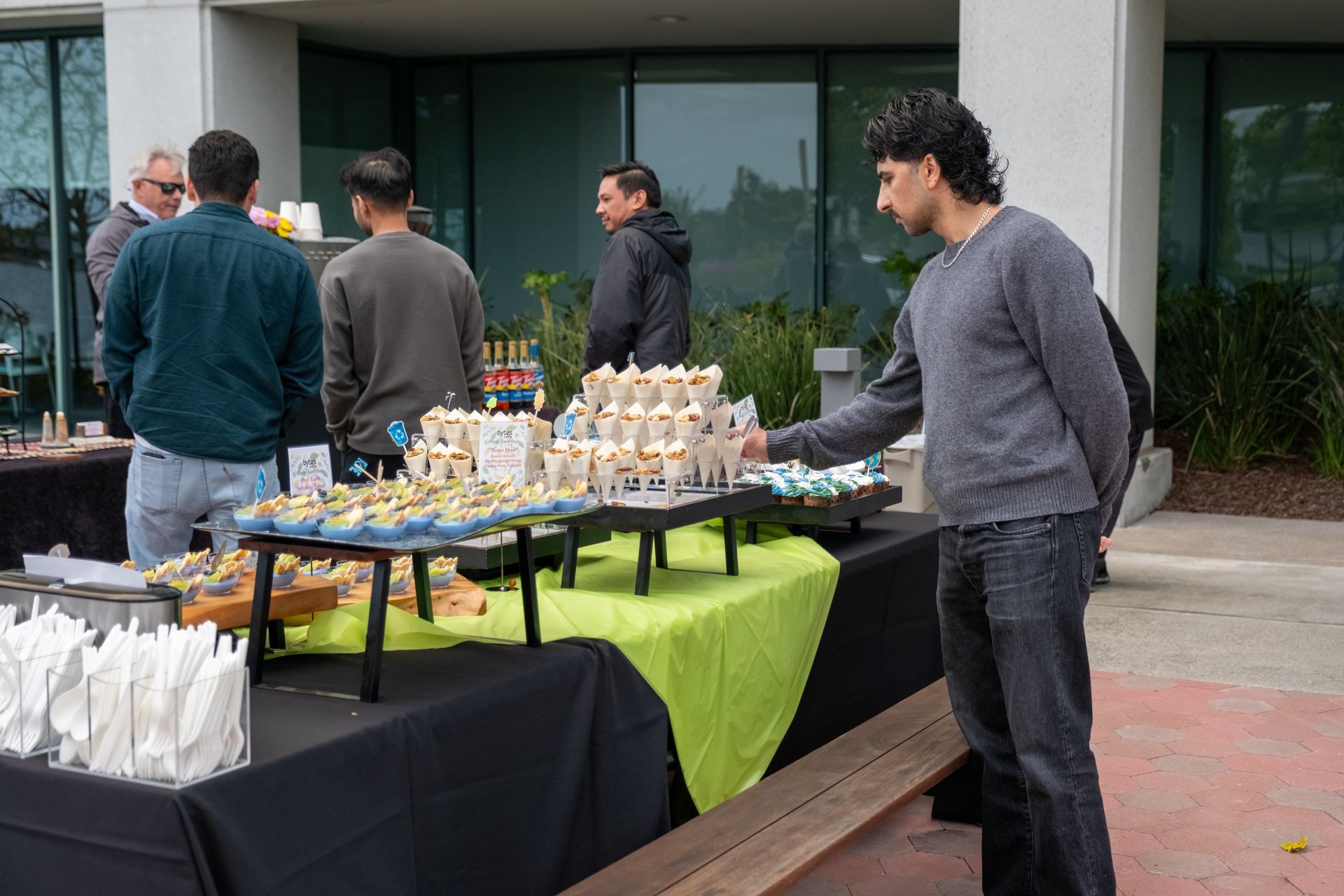 People at a food table, person reaching for a cupcake. Outdoor setting, other people in background.
