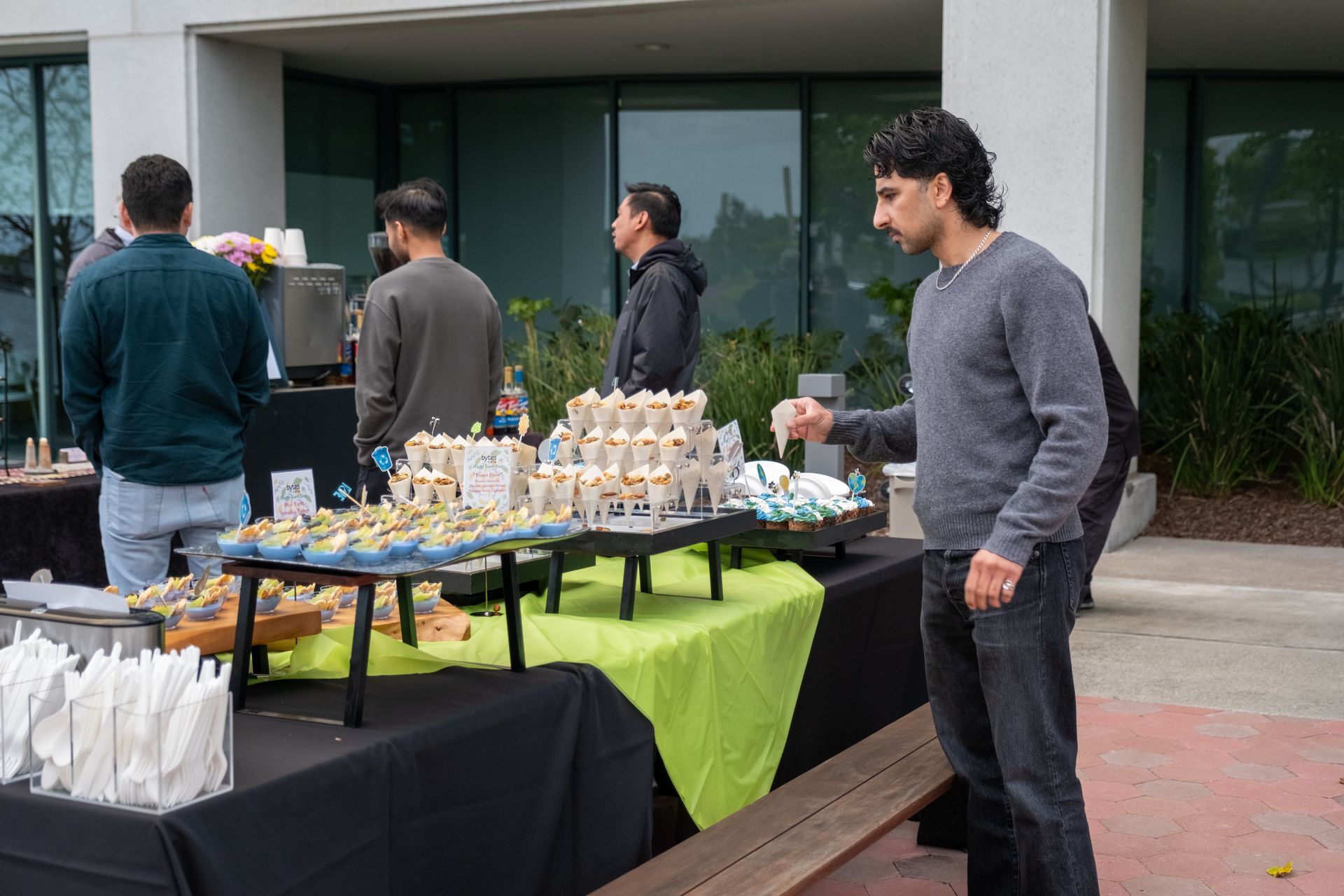 People at an outdoor event with food tables. A person reaches for a cupcake.