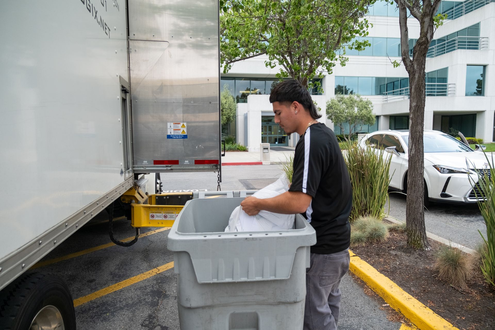 Man loading white bags into a gray bin from a delivery truck parked outside a building.