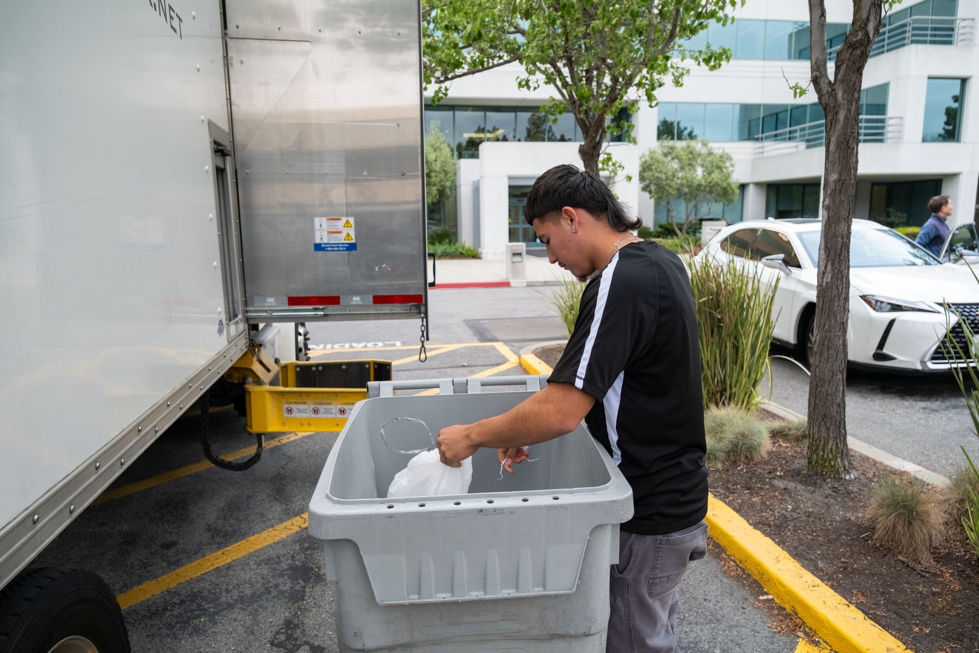 Person unloading items from a truck into a gray bin outdoors, near a building and parked cars.