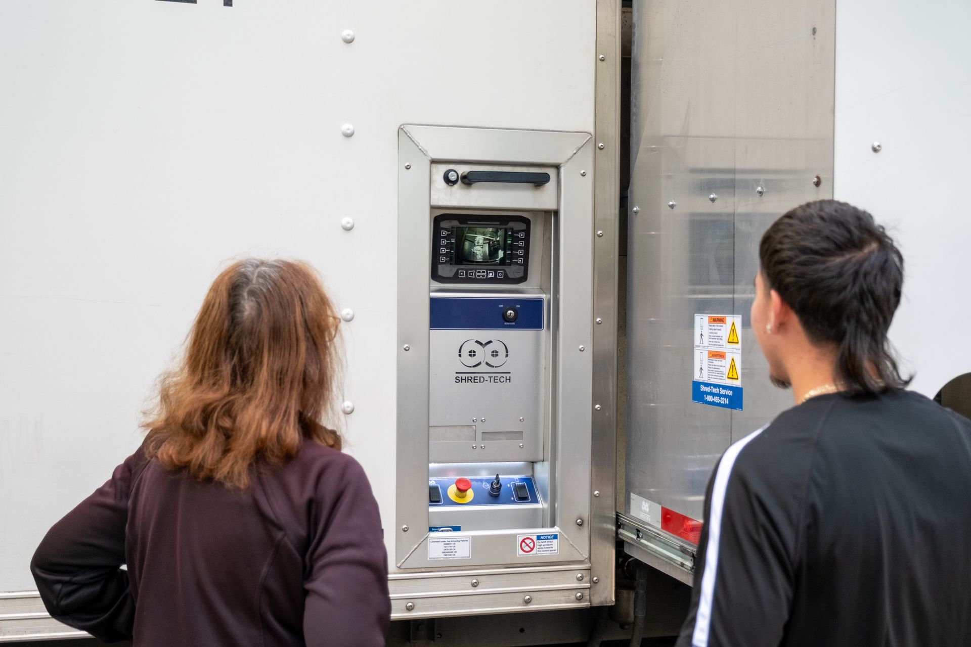 Two people looking at a control panel mounted on a truck. The panel has a screen and buttons.