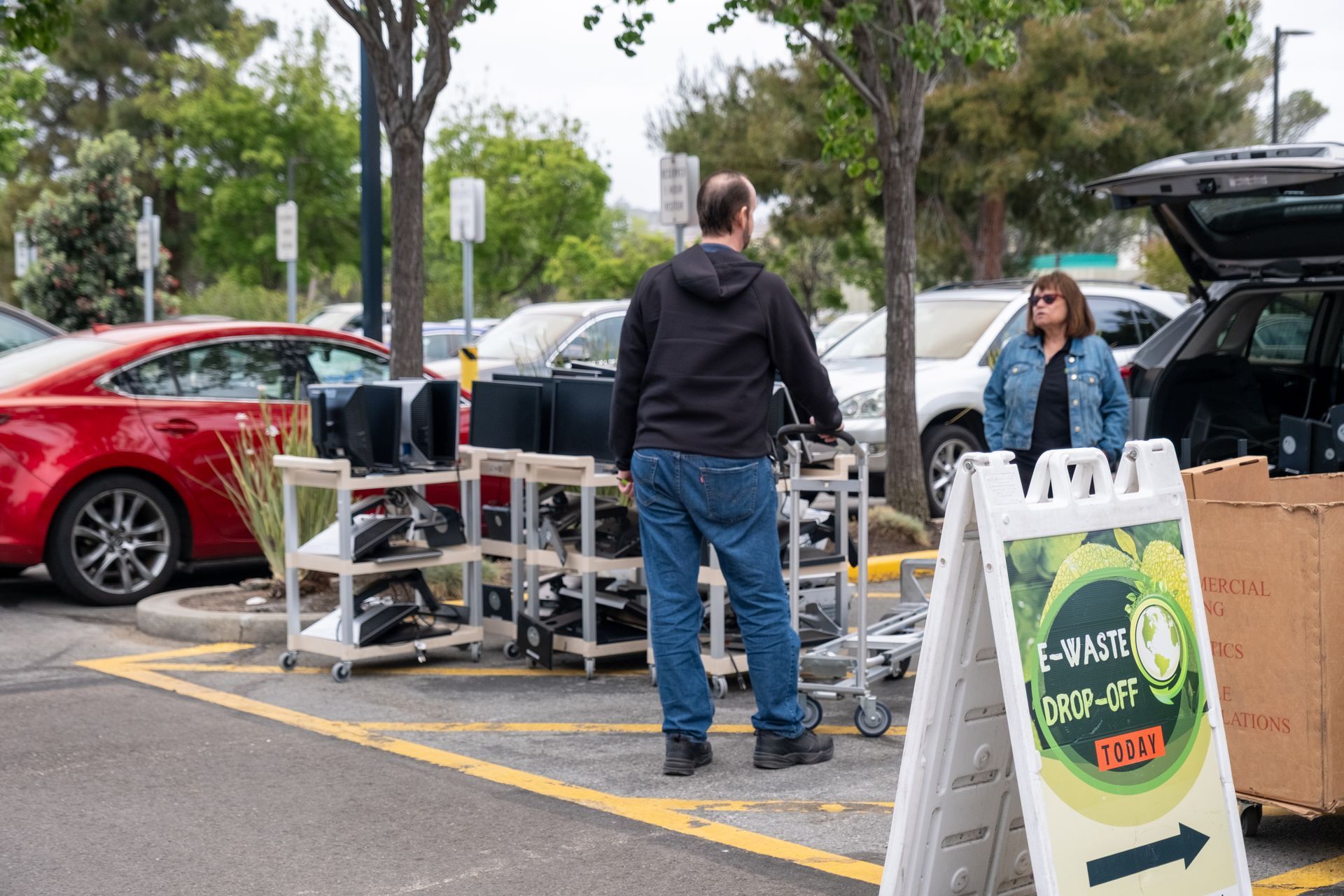 Man at e-waste drop-off, standing near a cart of electronics. Woman by an open car trunk. Parking lot setting.