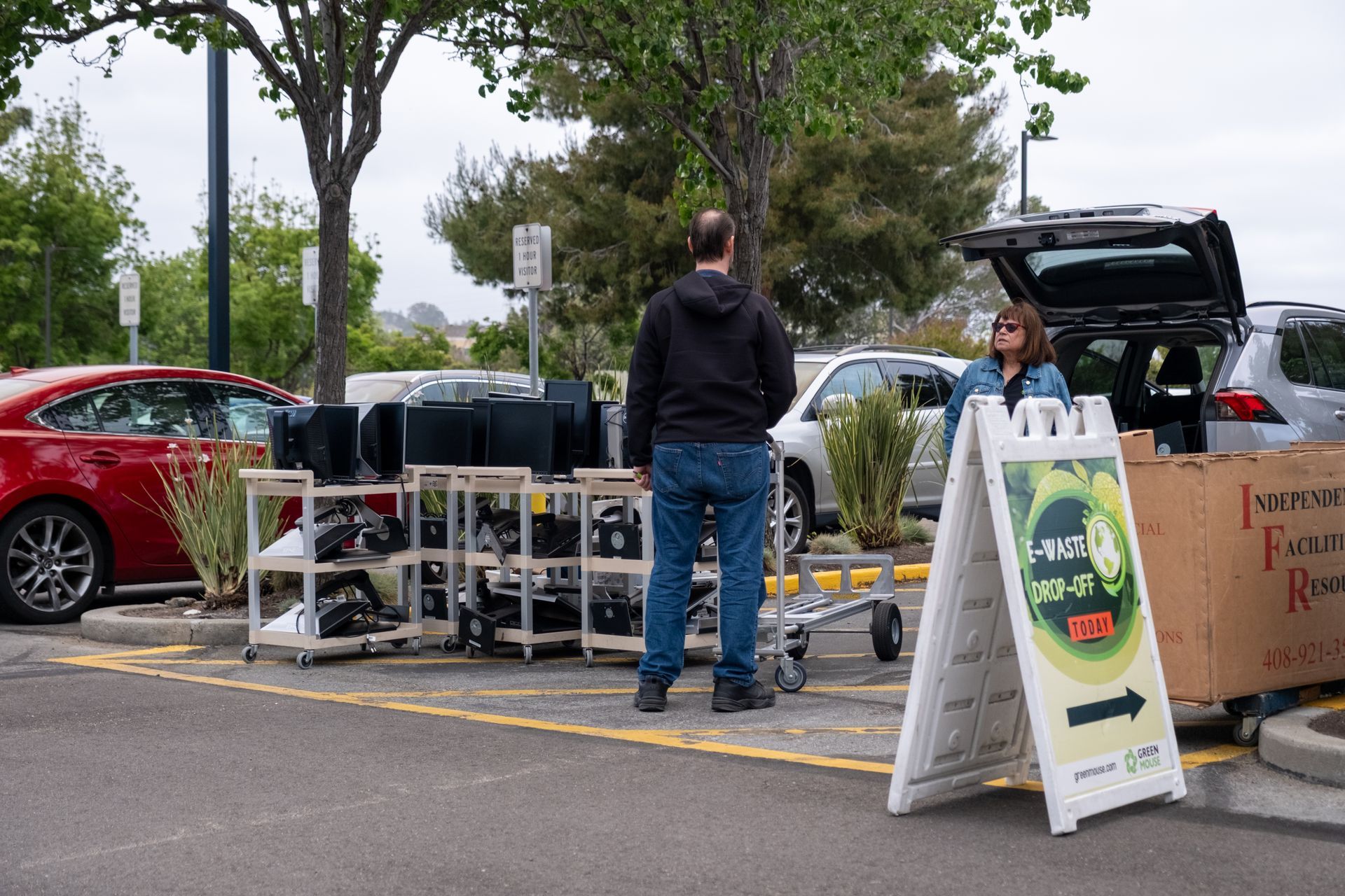 People selling electronics in a parking lot. A-frame sign and open car trunk visible.