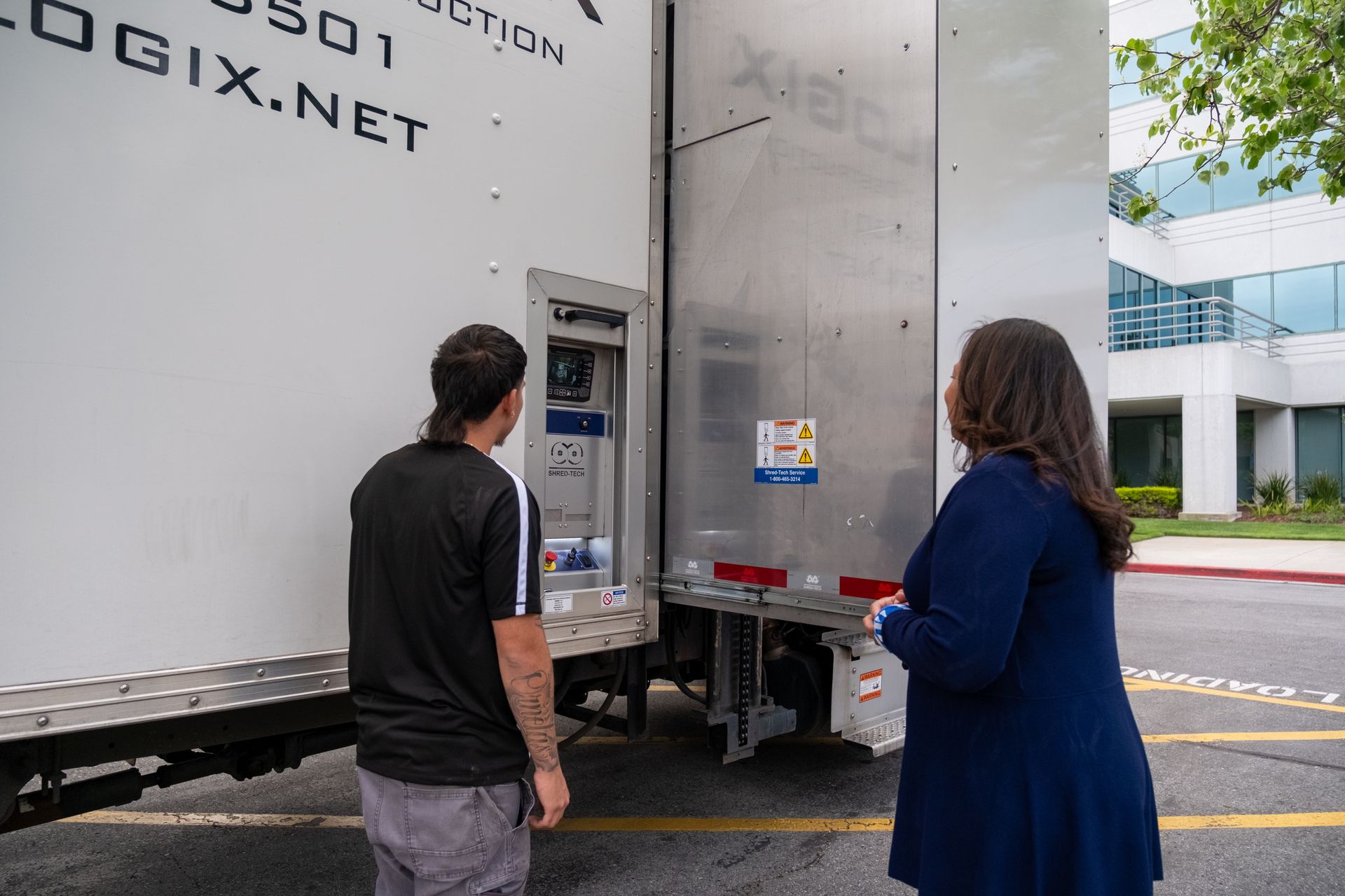 Two people examining the rear of a semi-truck with an open door. White truck, outdoors.