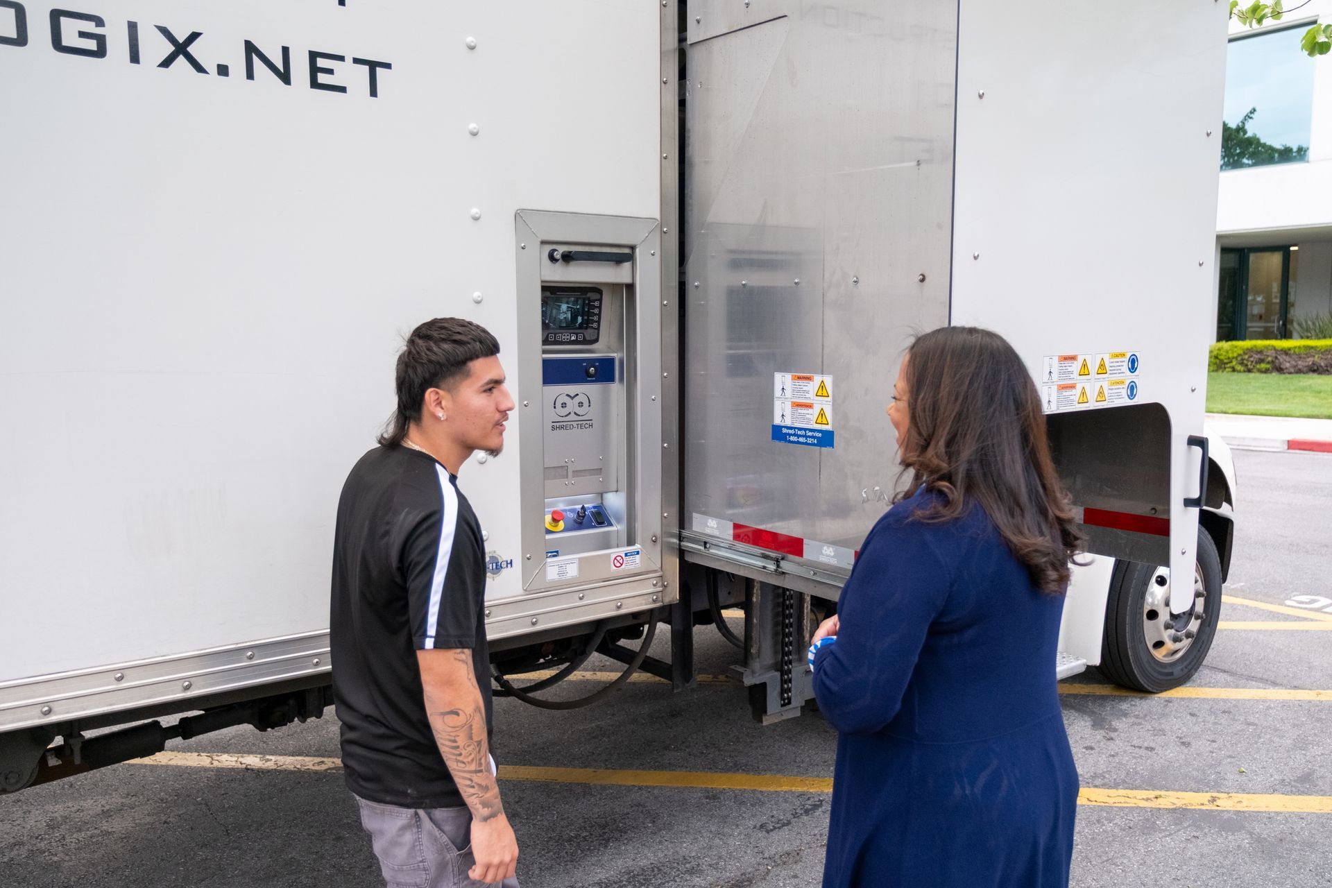 A person talking with another person next to a large white truck with the back door open outdoors.