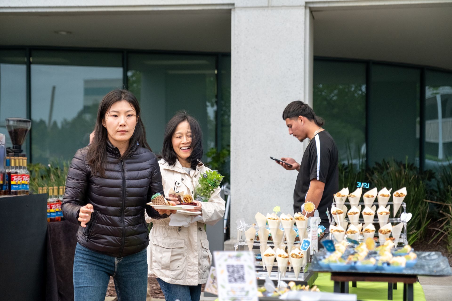 Two women holding food smile near a table of treats, a man on phone in the background. Outdoor setting.