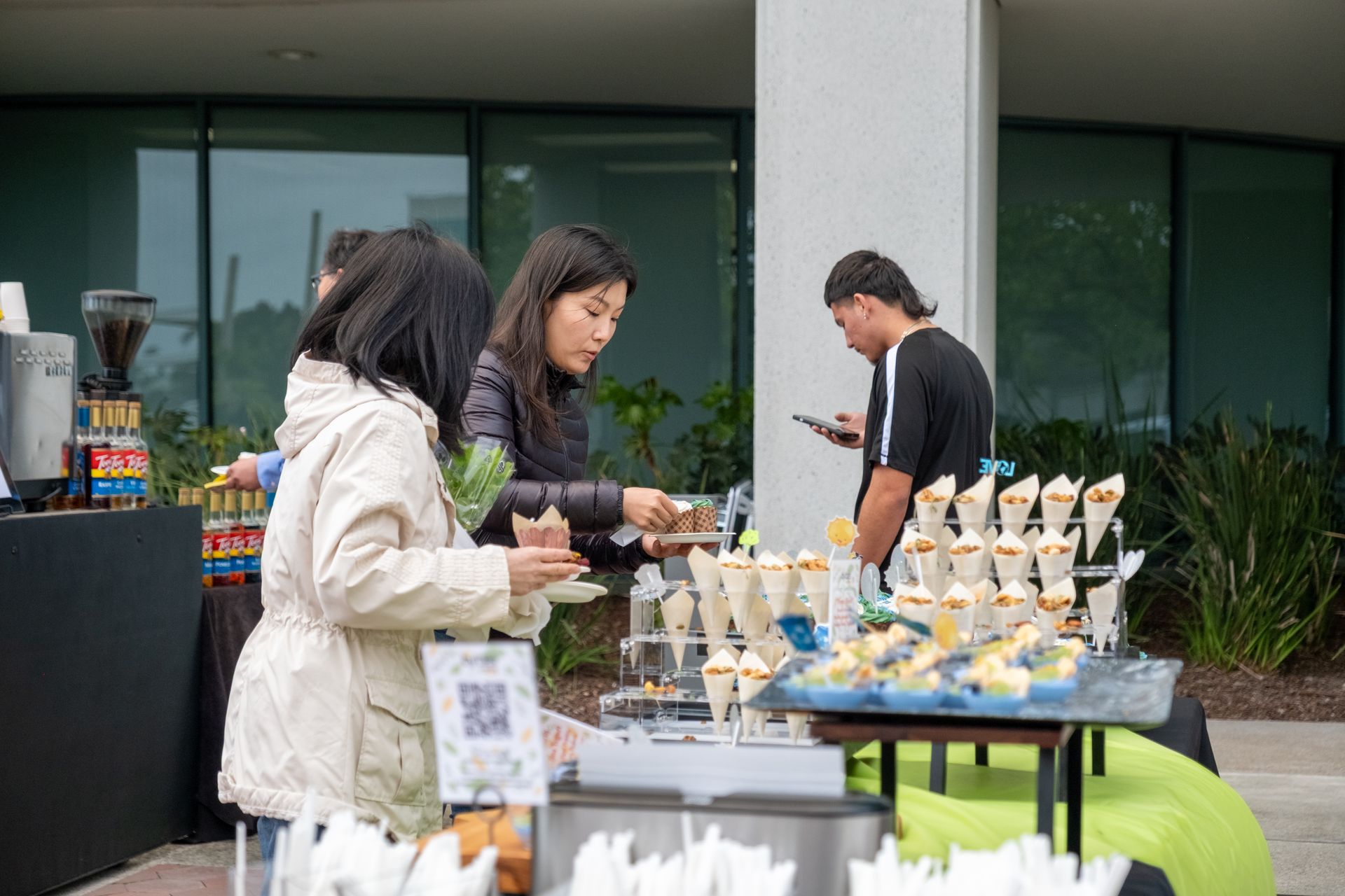 People at an outdoor food table selecting treats. The table has cones, and a person is taking a photo.