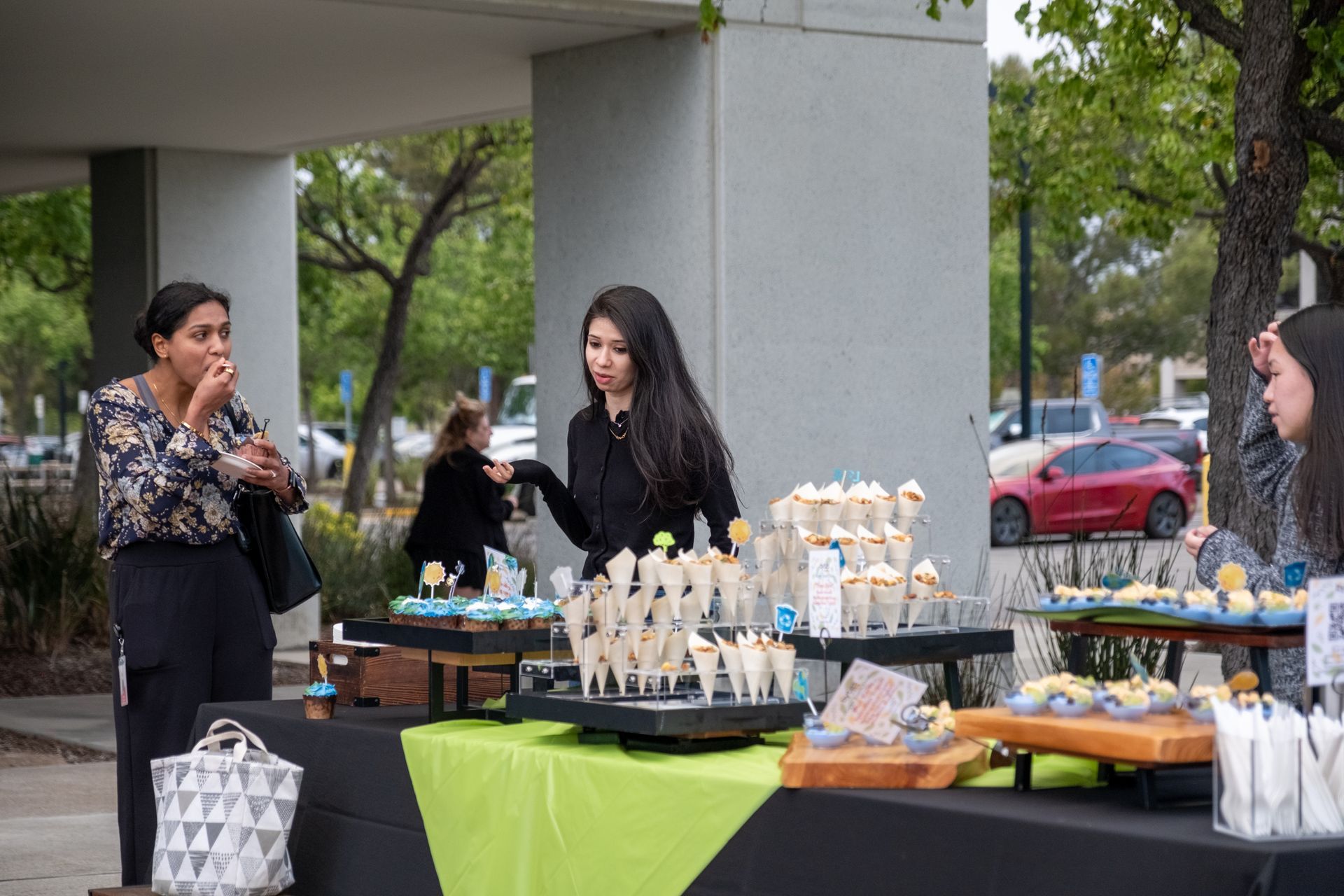 Women at a dessert table with cupcakes outside a building, one eating, others looking.