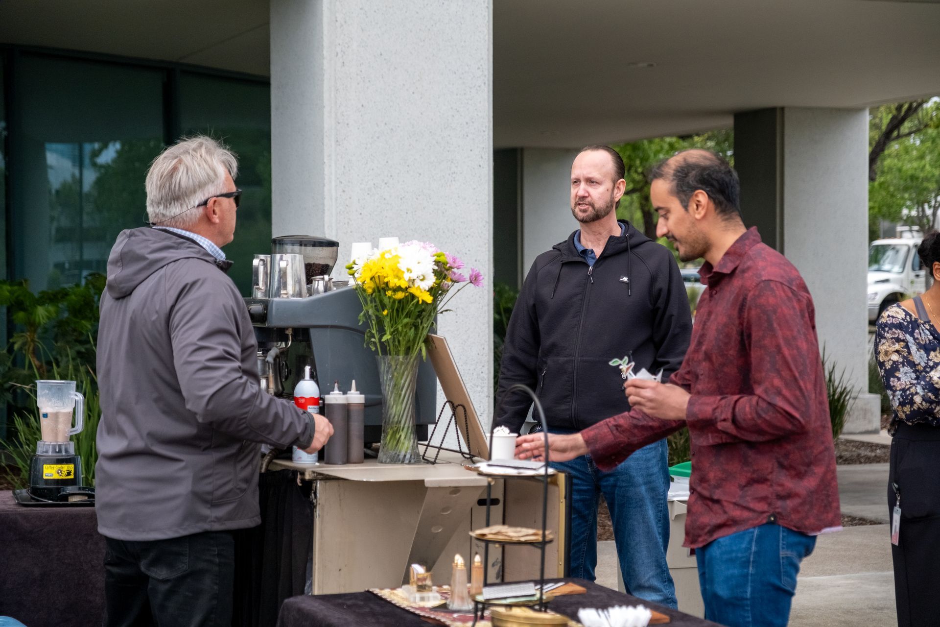 People at an outdoor coffee station. A man serves drinks, and two others watch. Flowers and coffee supplies are on the table.
