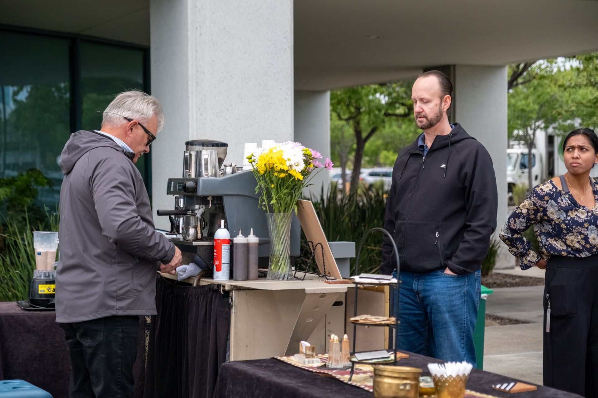 Outdoor coffee stand with people. Man prepares coffee, two customers watch. Flowers and pastries on the table.