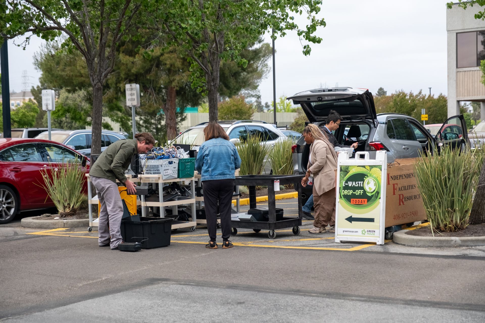People setting up a table with plants in a parking lot next to cars; sign reads 