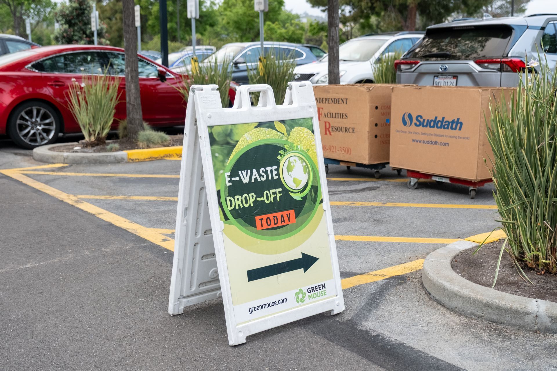 White A-frame sign promoting E-waste drop-off in parking lot, with arrow pointing right.