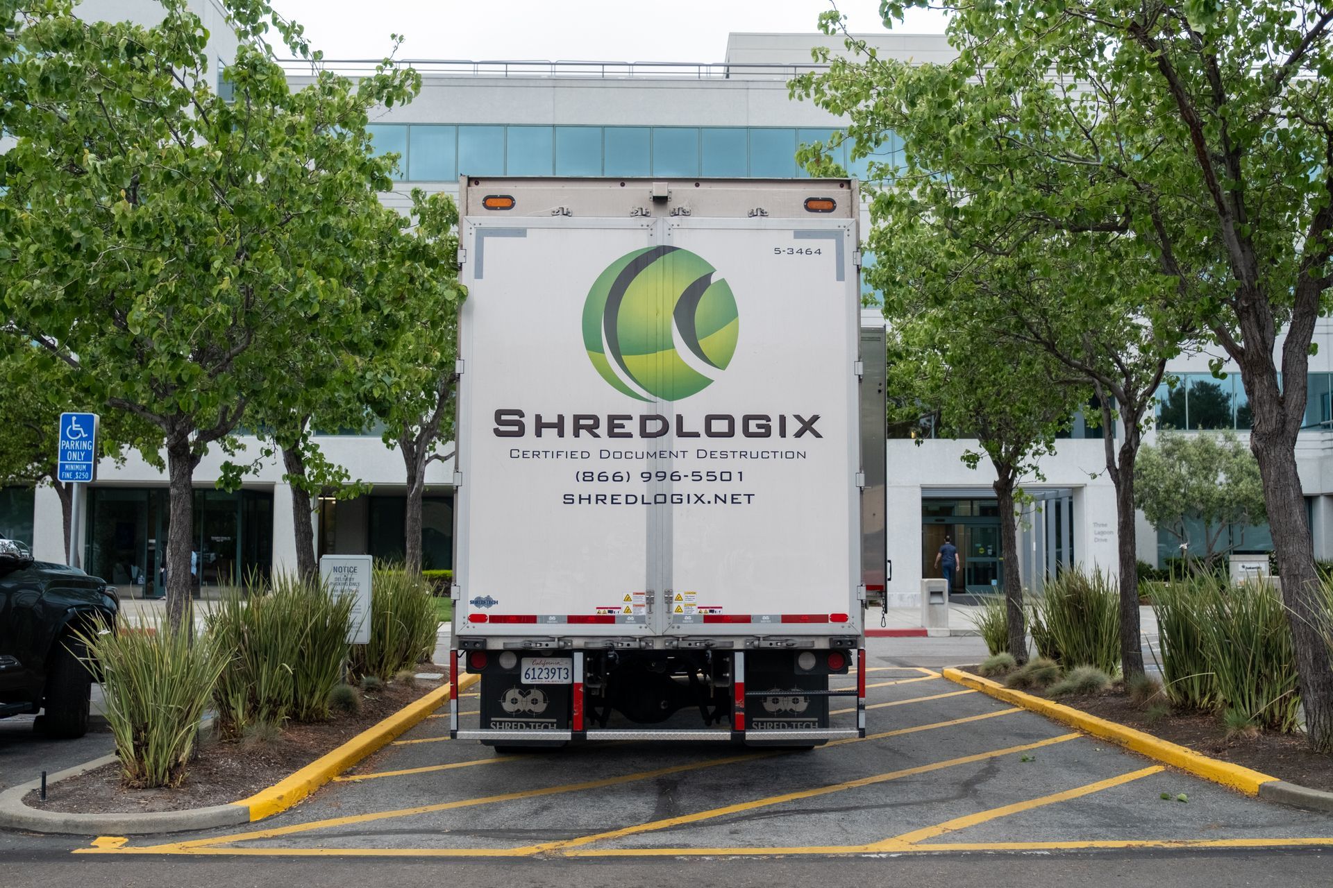 A white Shredlogix truck parked in front of a building; trees and bushes flank it.