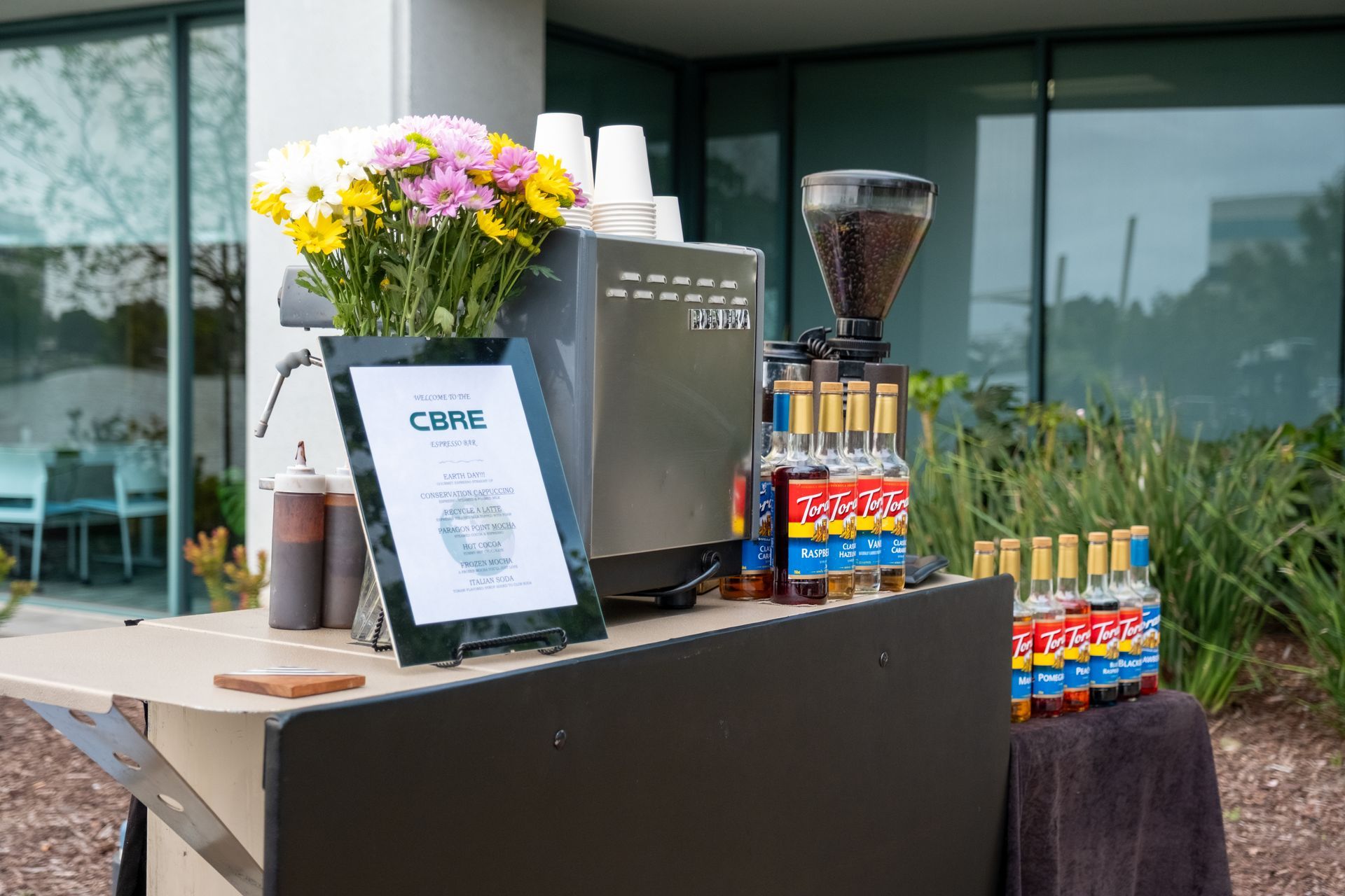Coffee cart with coffee maker, syrups, flowers, and sign outdoors.
