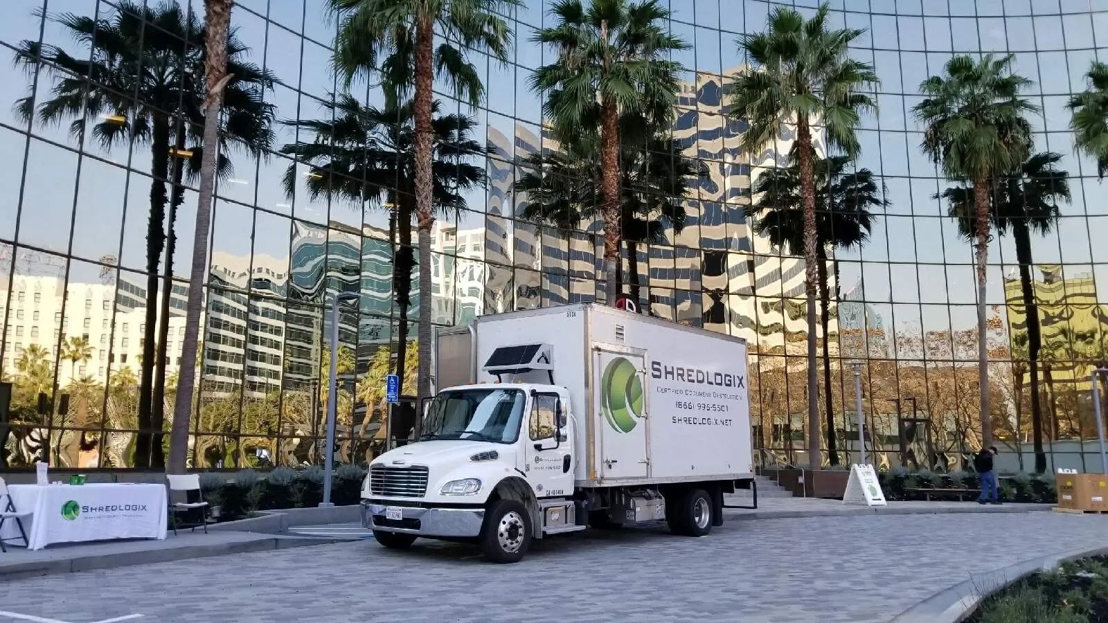 White delivery truck with logo parked outside glass building with palm trees.