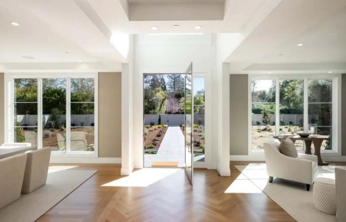 A modern, bright entryway with open glass door and exterior view, flanked by white-framed windows and neutral-toned walls.