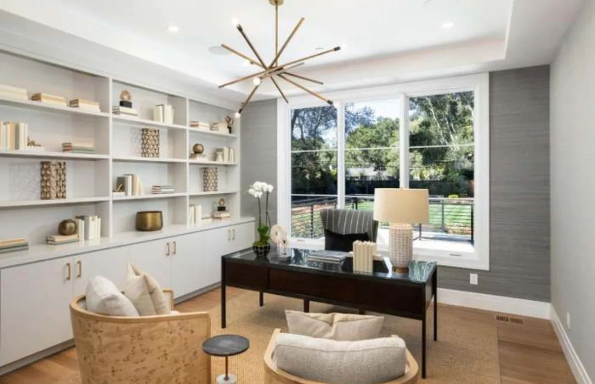 Home office with built-in white shelves, a desk, chairs, and a window overlooking greenery.