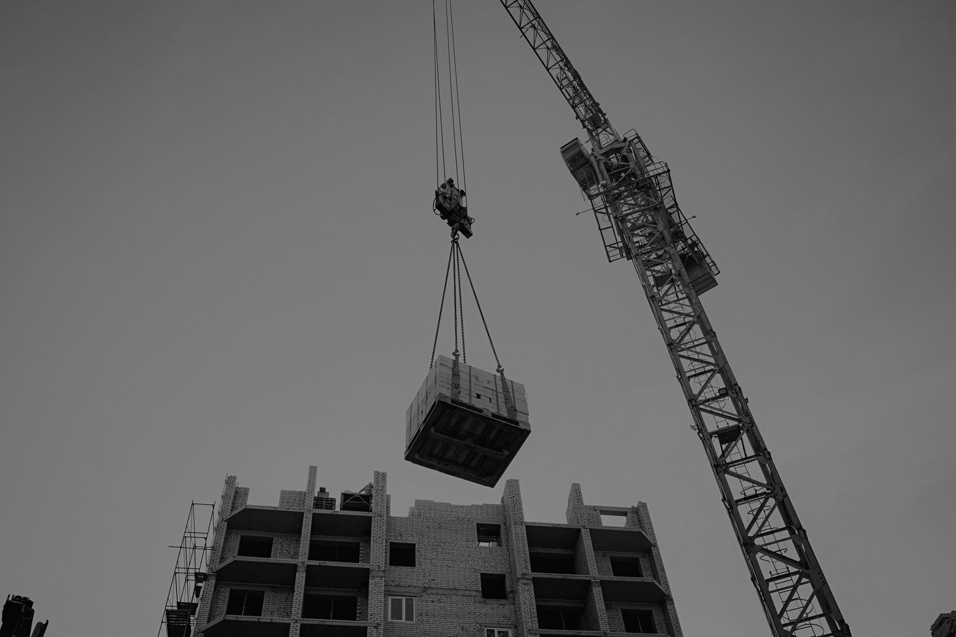 A black and white photo of a crane lifting a container over a building under construction.