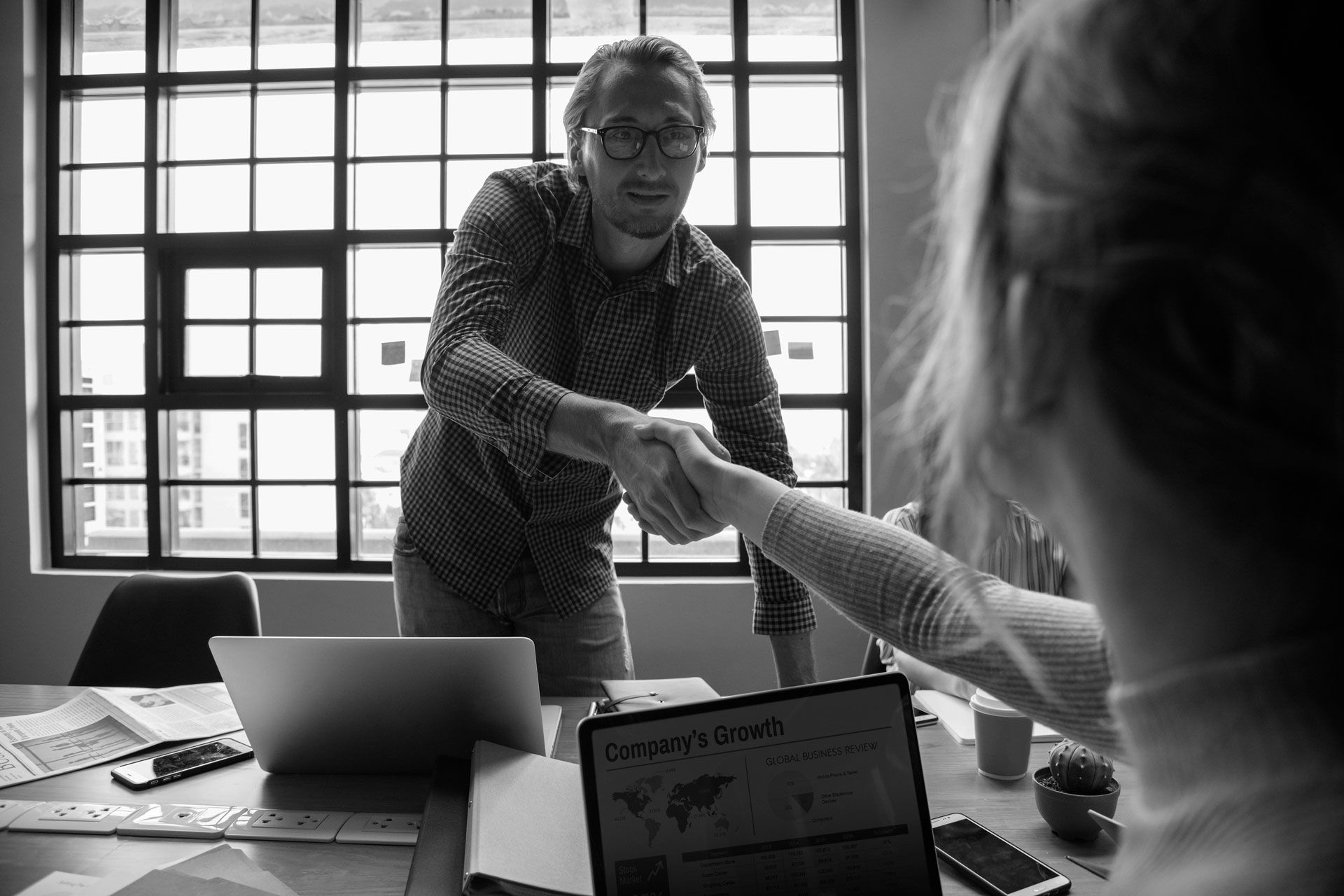 Man in patterned shirt shaking hands with a person, in an office with a window and laptops.
