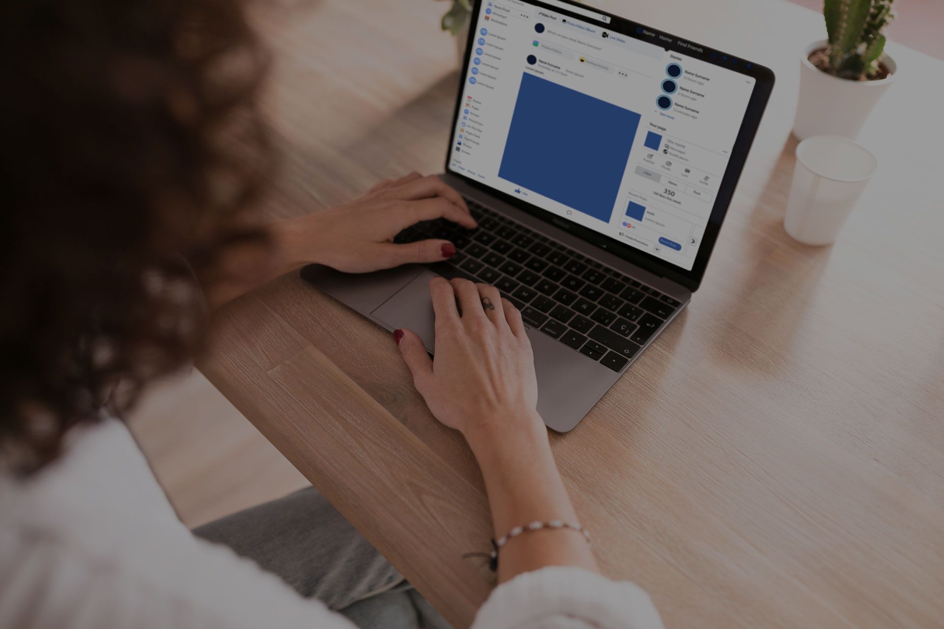 Person typing on a laptop at a wooden desk, screen showing a social media feed.