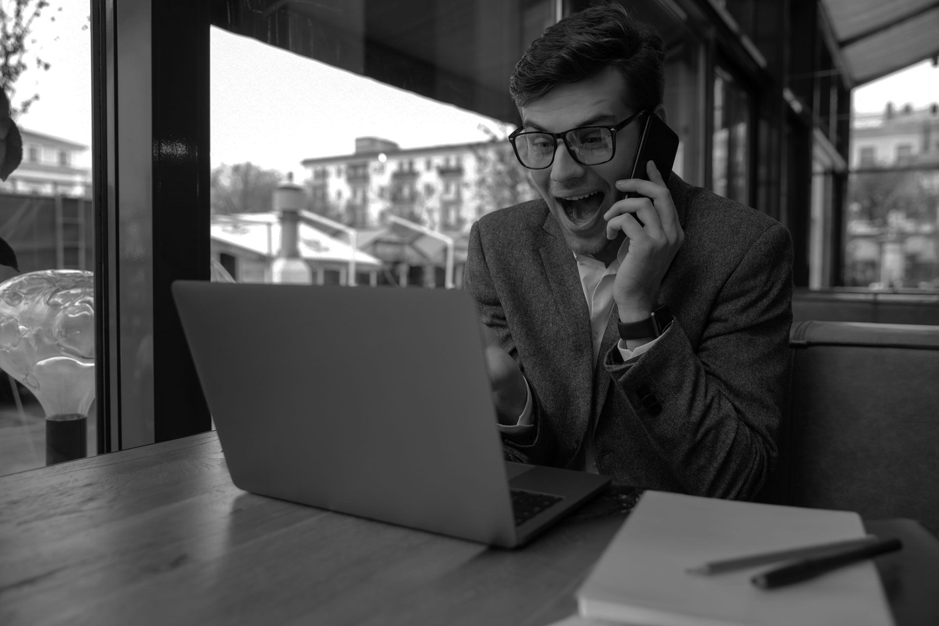 Man with glasses, laptop, and phone, surprised expression. Sitting at a table indoors.
