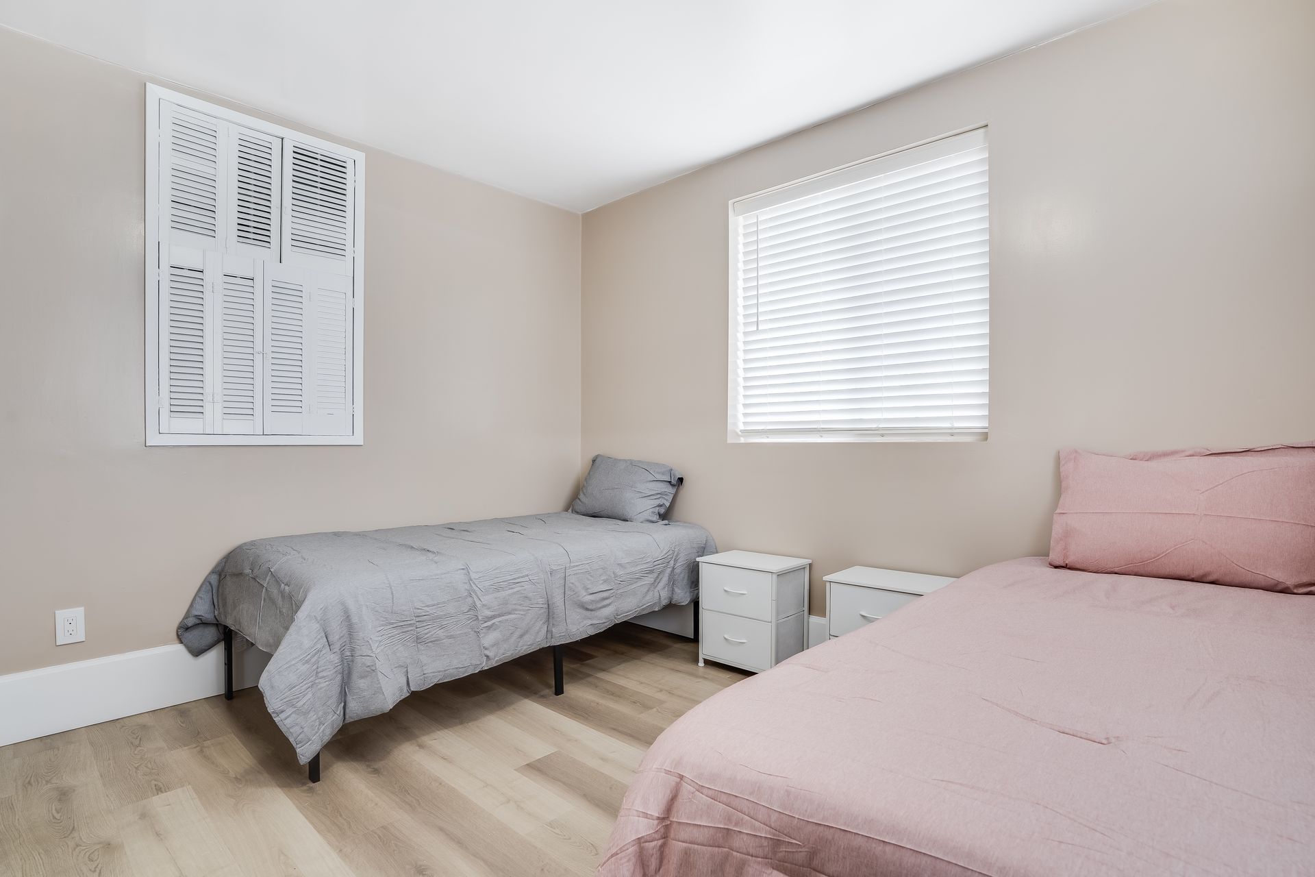 Two twin beds in a bedroom, one gray and one pink, with matching nightstands. Beige walls, a window, and art.