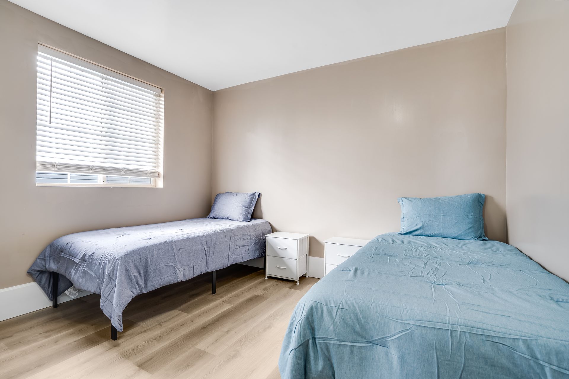 Two twin beds in a room with beige walls, a window with blinds, and a small white nightstand.