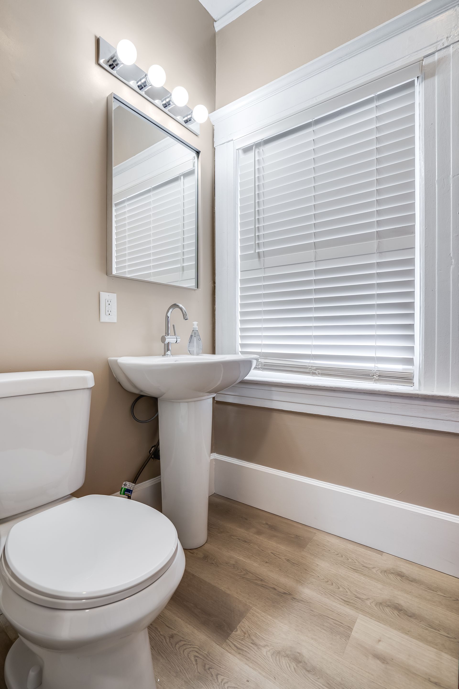Small bathroom with a toilet, pedestal sink, mirror, and window with blinds; beige walls, wooden floor.