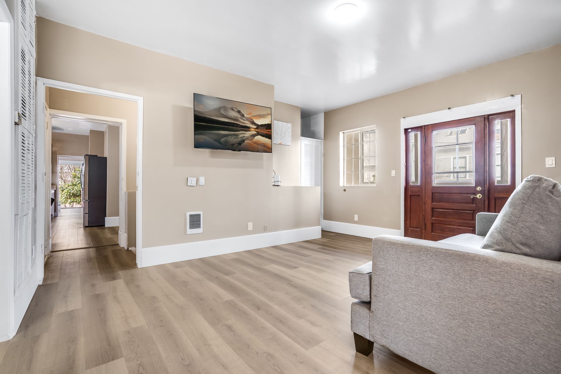 Living room with a light gray couch, TV, and brown hardwood door. Beige walls, wood floors.
