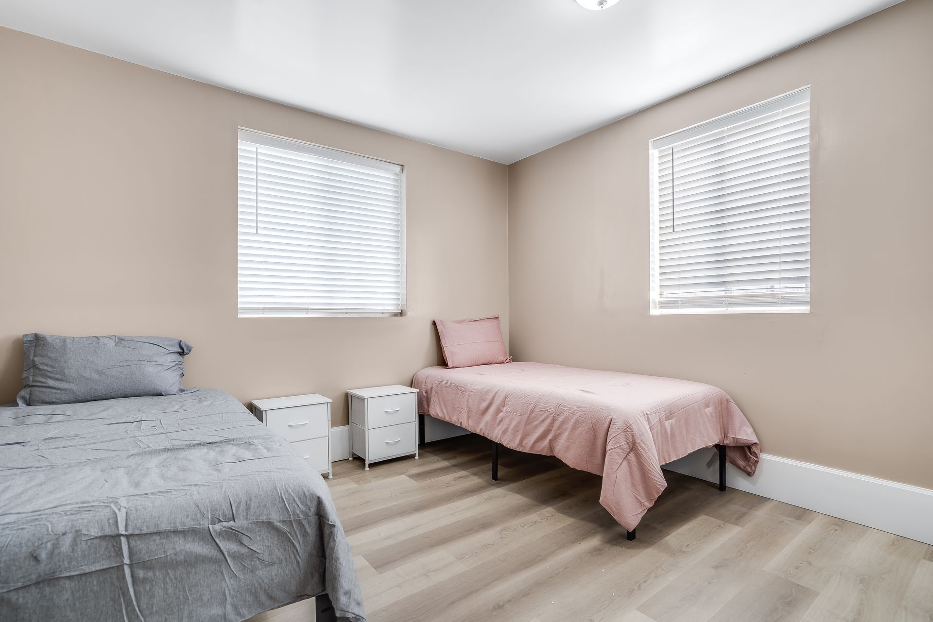 Two twin beds in a room with tan walls, two windows with blinds, and light wood floors.